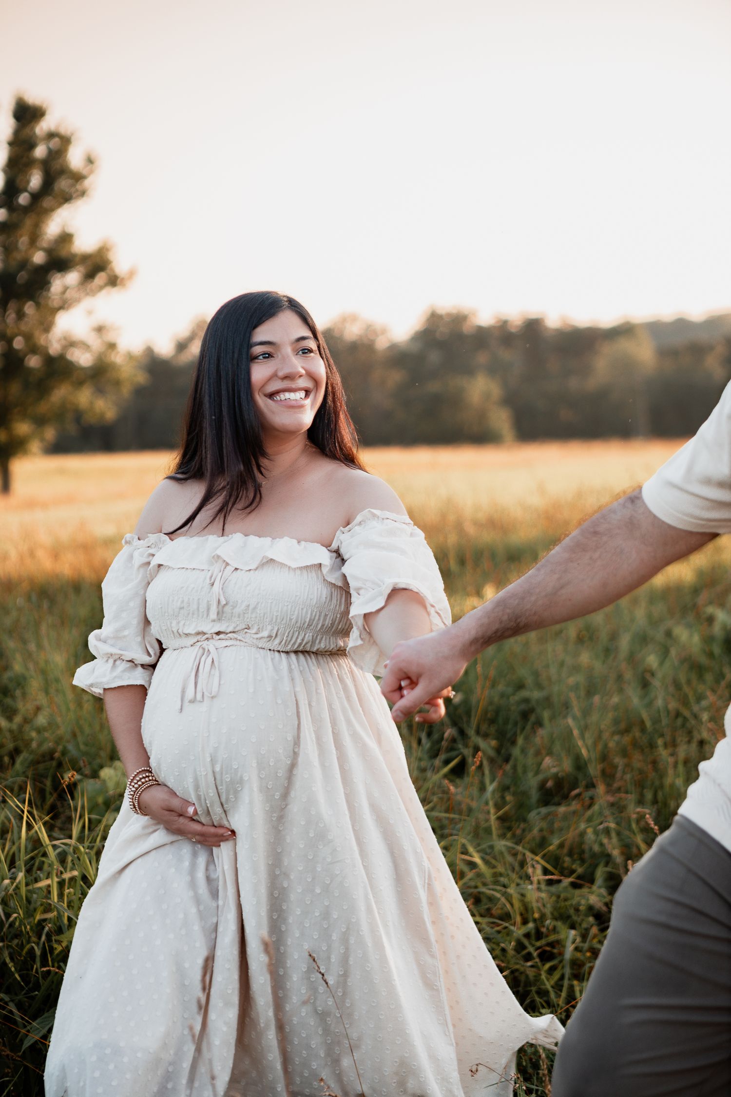A golden hour maternity photo in a field shows an expecting mother in a flowing off-shoulder cream dress.