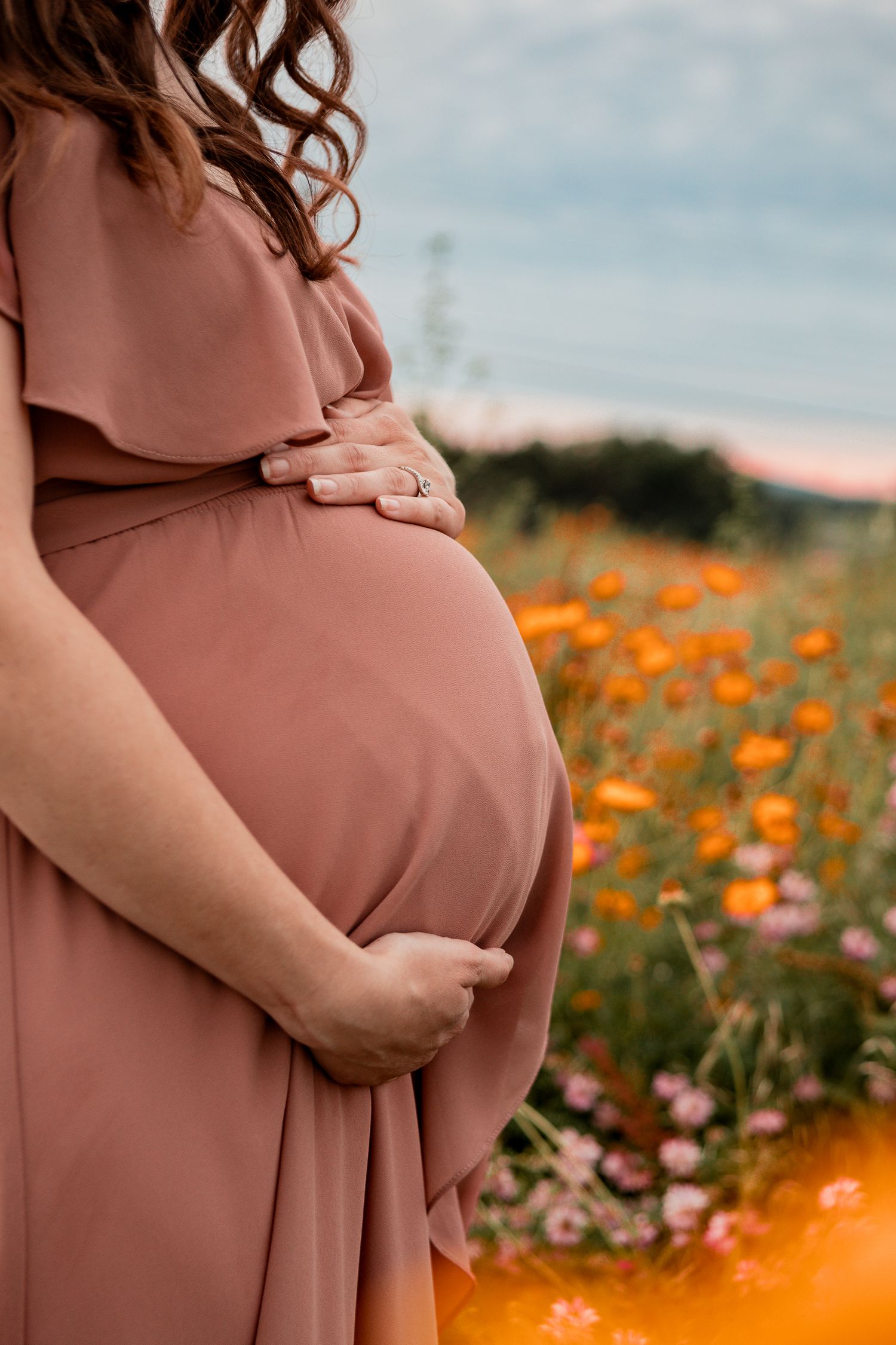 Close-up of hands cradling a baby bump in a pink maxi dress among blooming wildflowers.