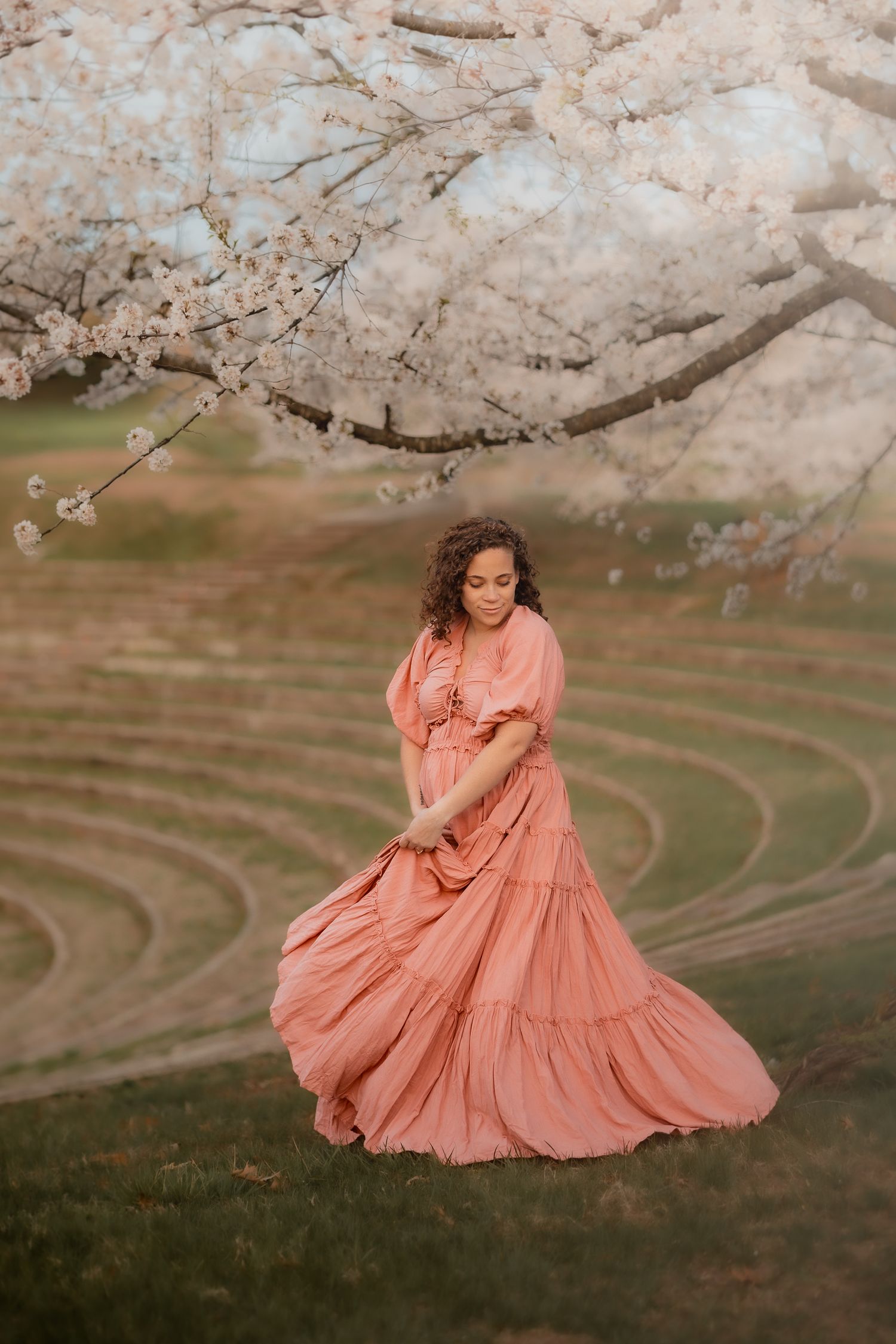 Woman in flowing pink dress twirling under blooming cherry blossom trees in a serene outdoor setting.