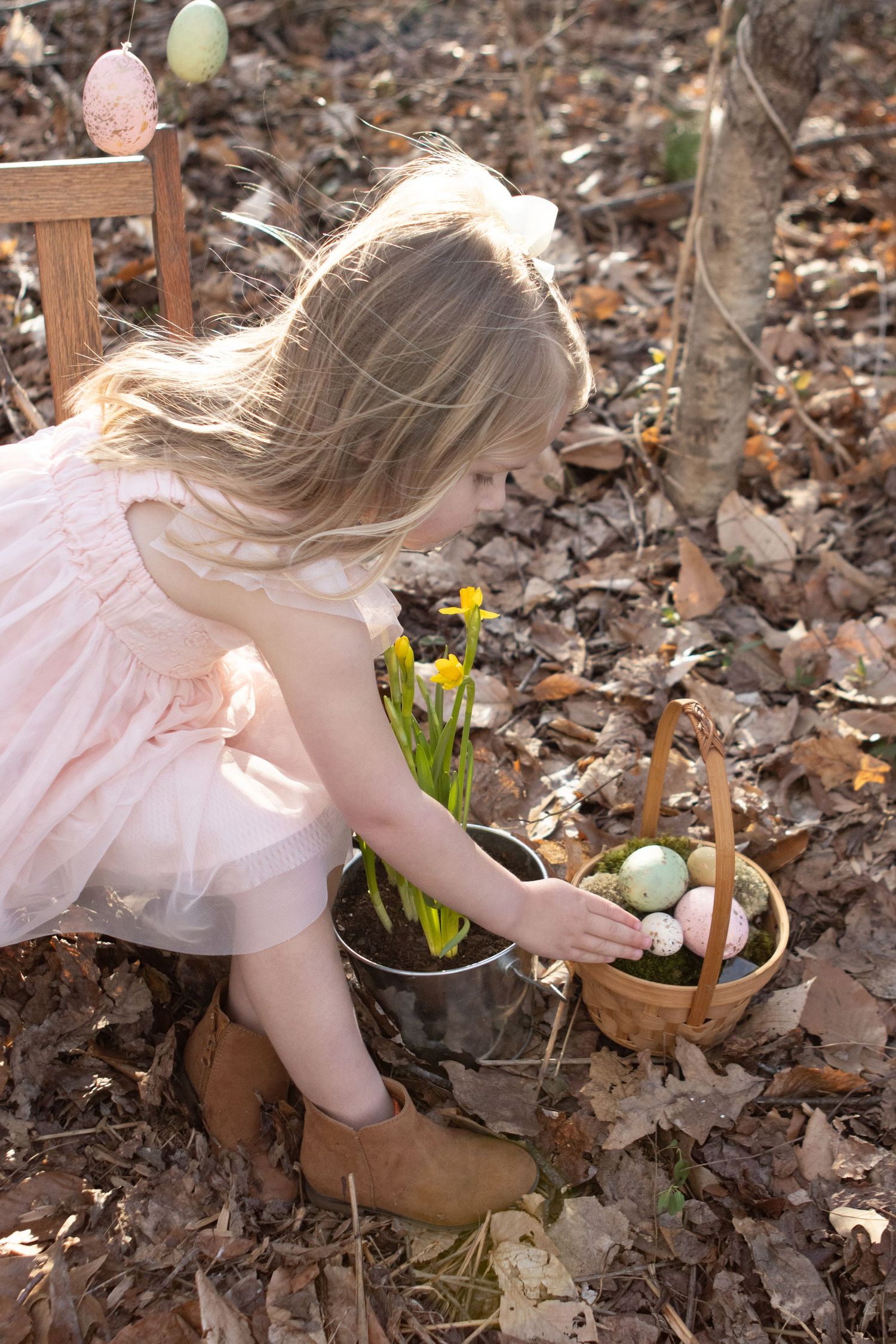 Child in pink dress reaching for Easter eggs in basket near blooming daffodils.
