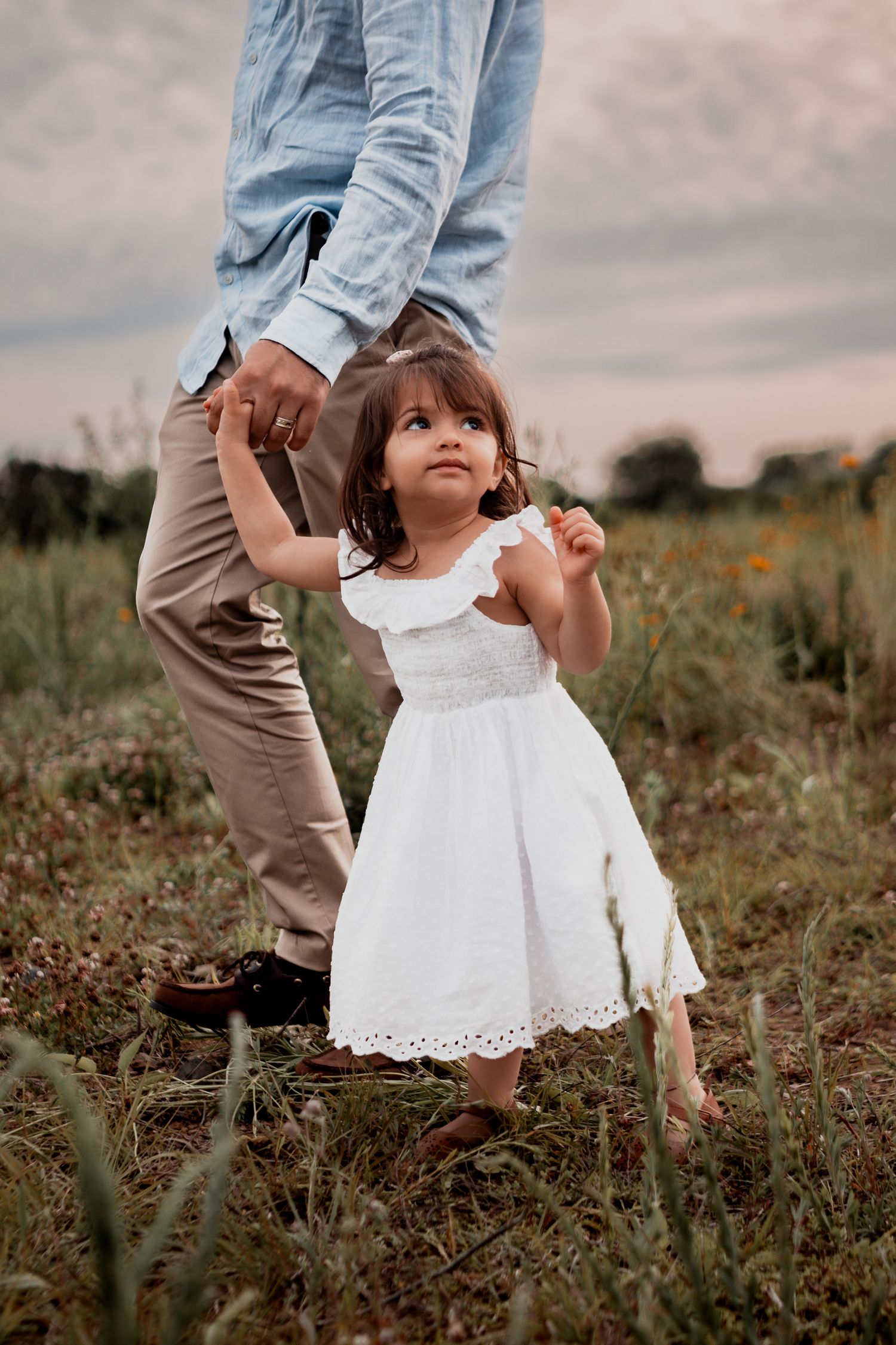 A small child in a white eyelet dress holds hands with someone in jeans and khakis while standing in a wildflower field.