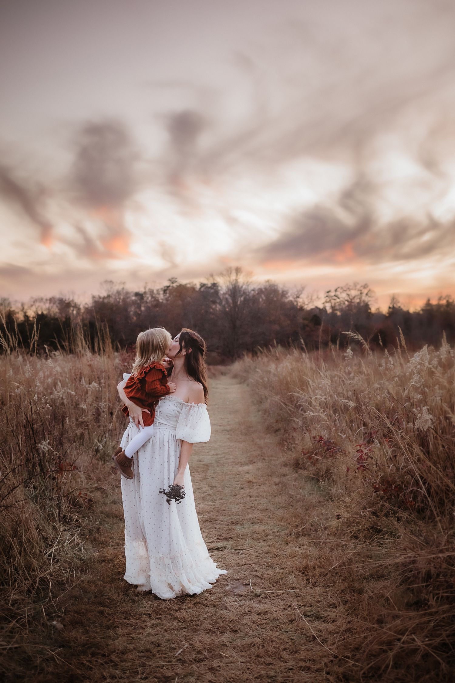 Two figures in white dresses dance together on a dirt path at sunset surrounded by tall grass and moody skies.