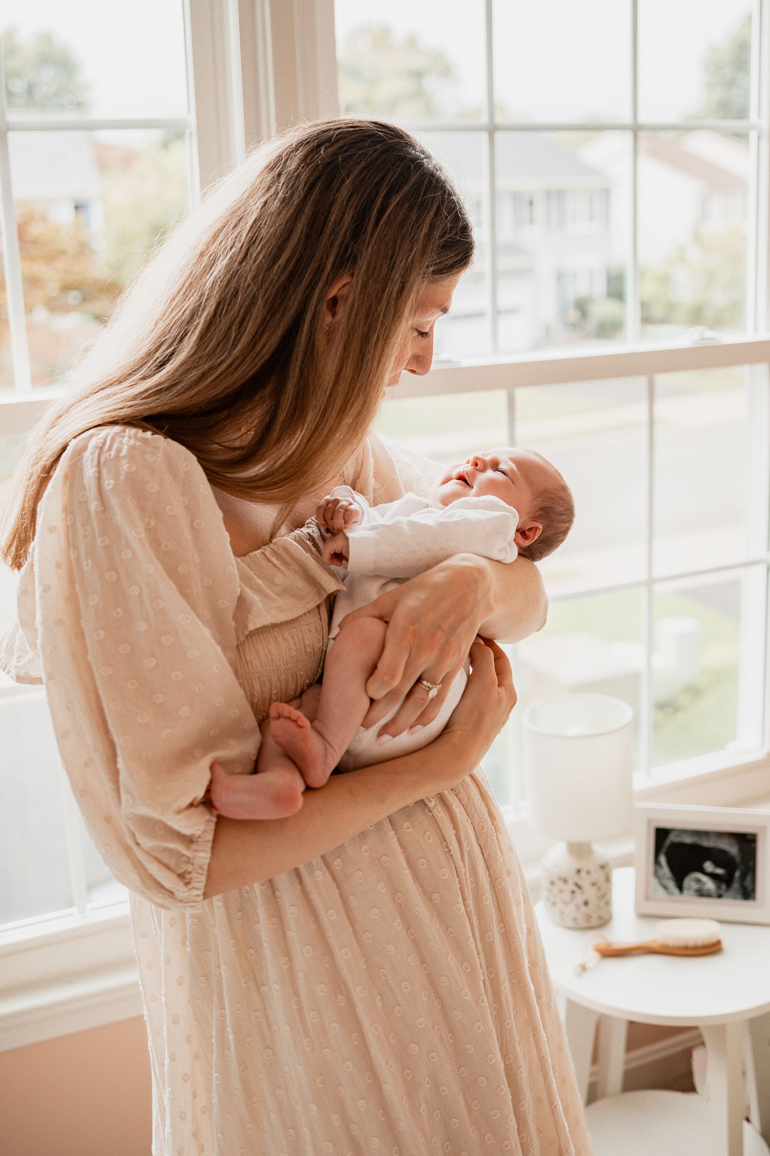 A woman in a beige dress cradles and lovingly interacts with a baby by a sunlit window.