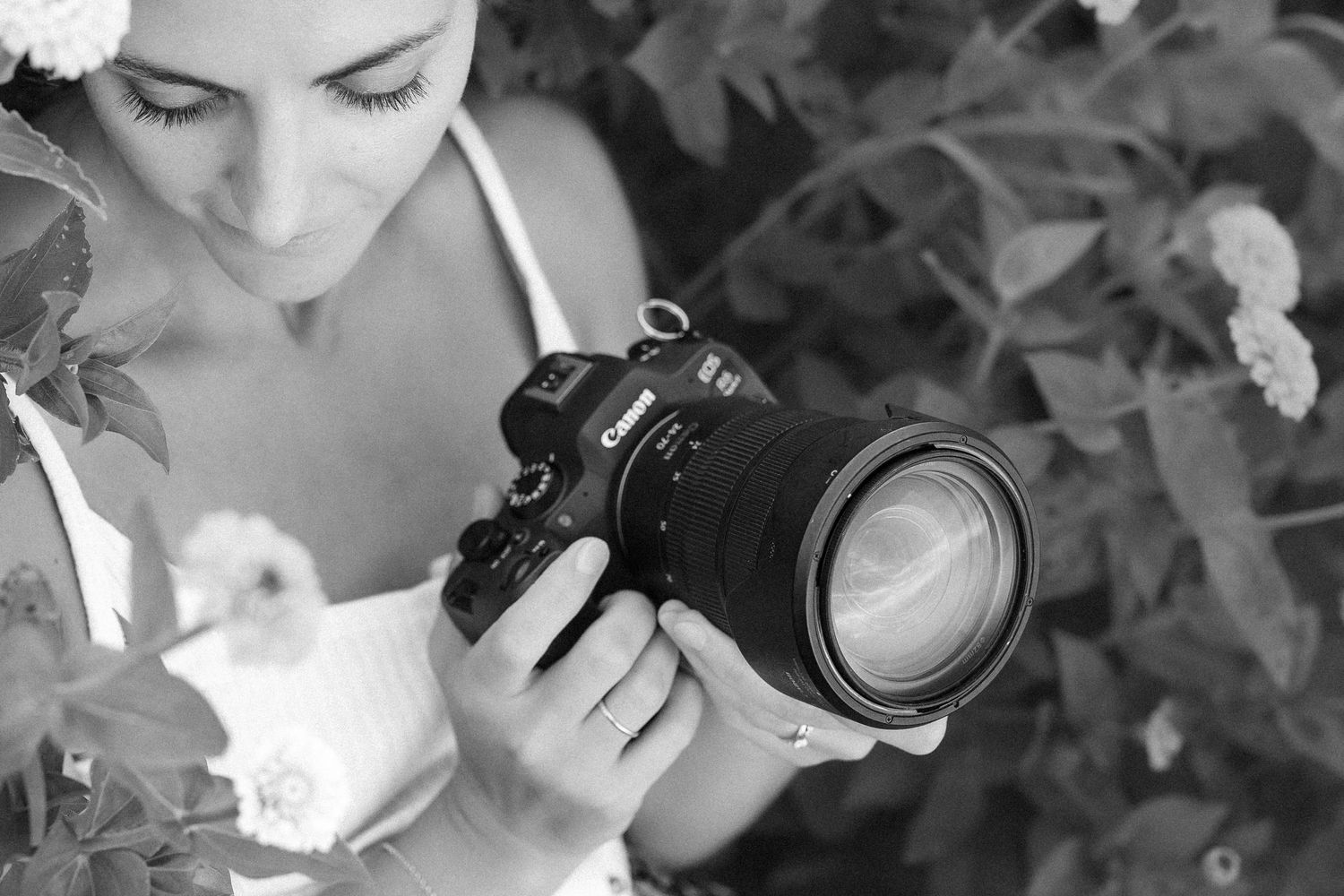 Black and white photo of a woman holding a DSLR camera outdoors surrounded by lush flowering plants.