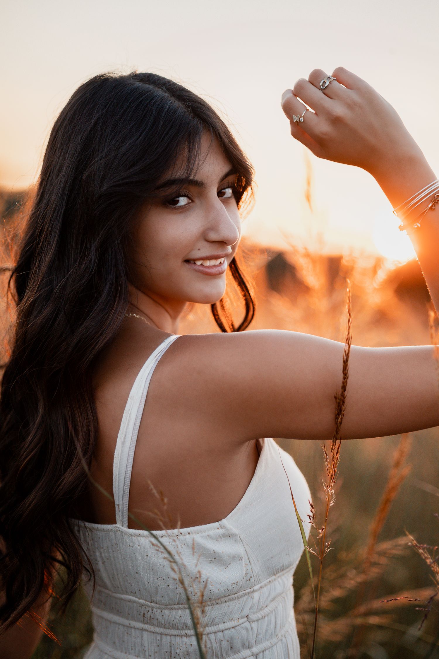 Person in white dress reaches toward sunset while standing in golden field.