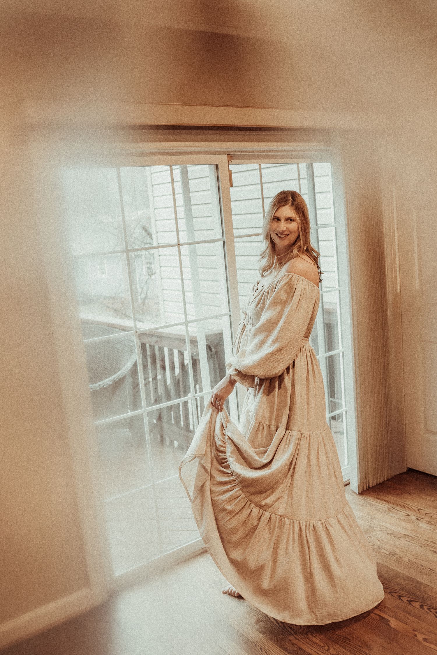 A figure in a flowing beige dress twirls gracefully in a sunlit room with wooden floors and large windows.