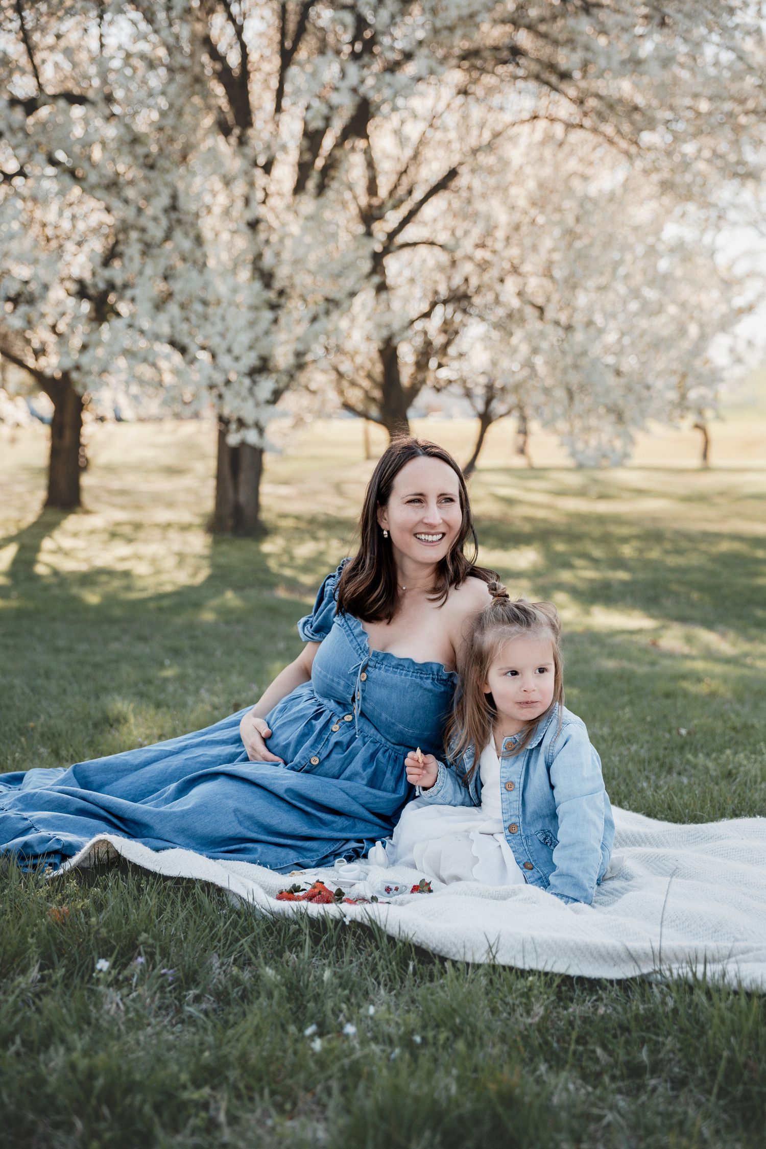 A smiling woman in a blue dress sits on a blanket with a young girl under blooming white trees.