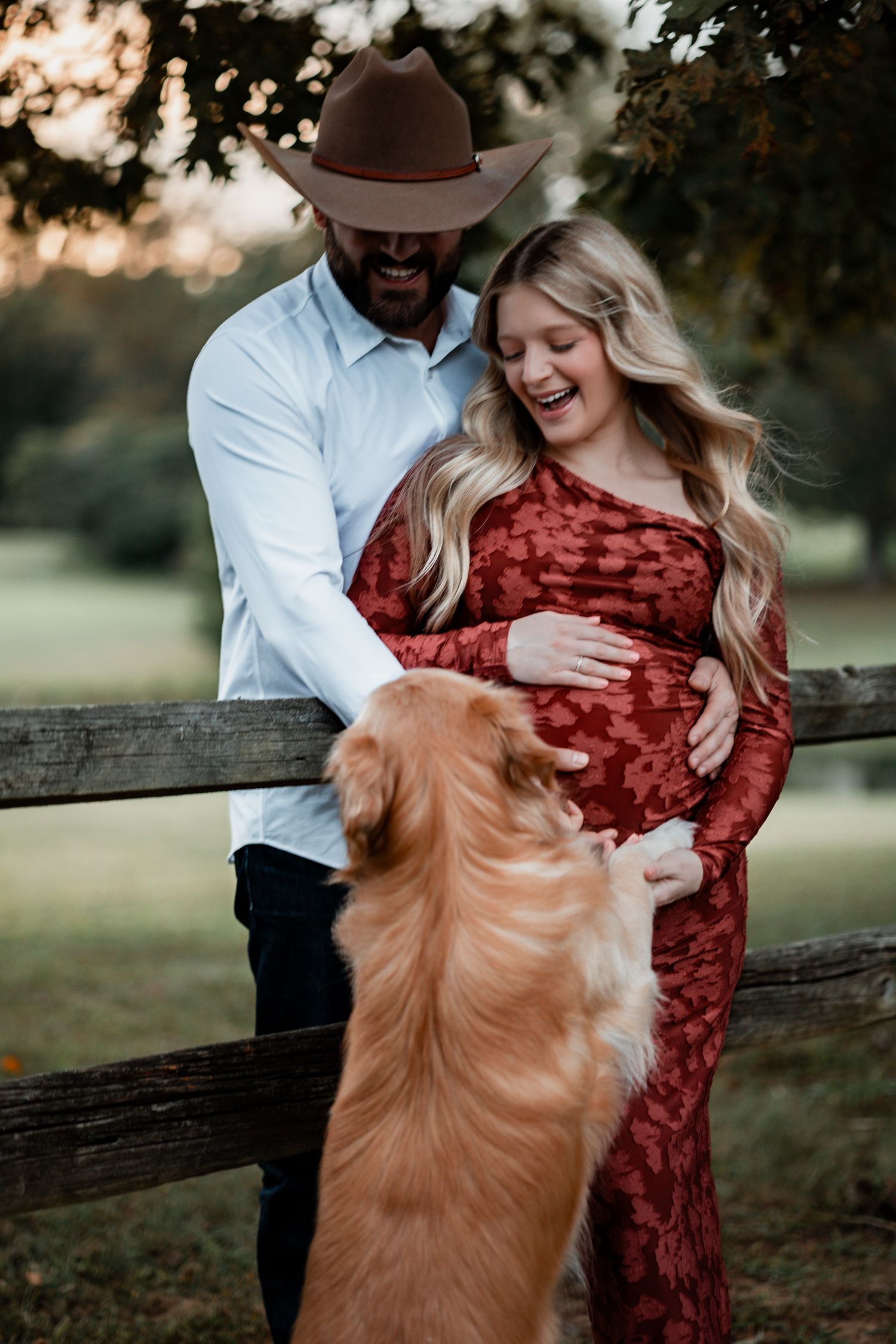 A couple and their golden retriever share a tender moment by a wooden fence at sunset.