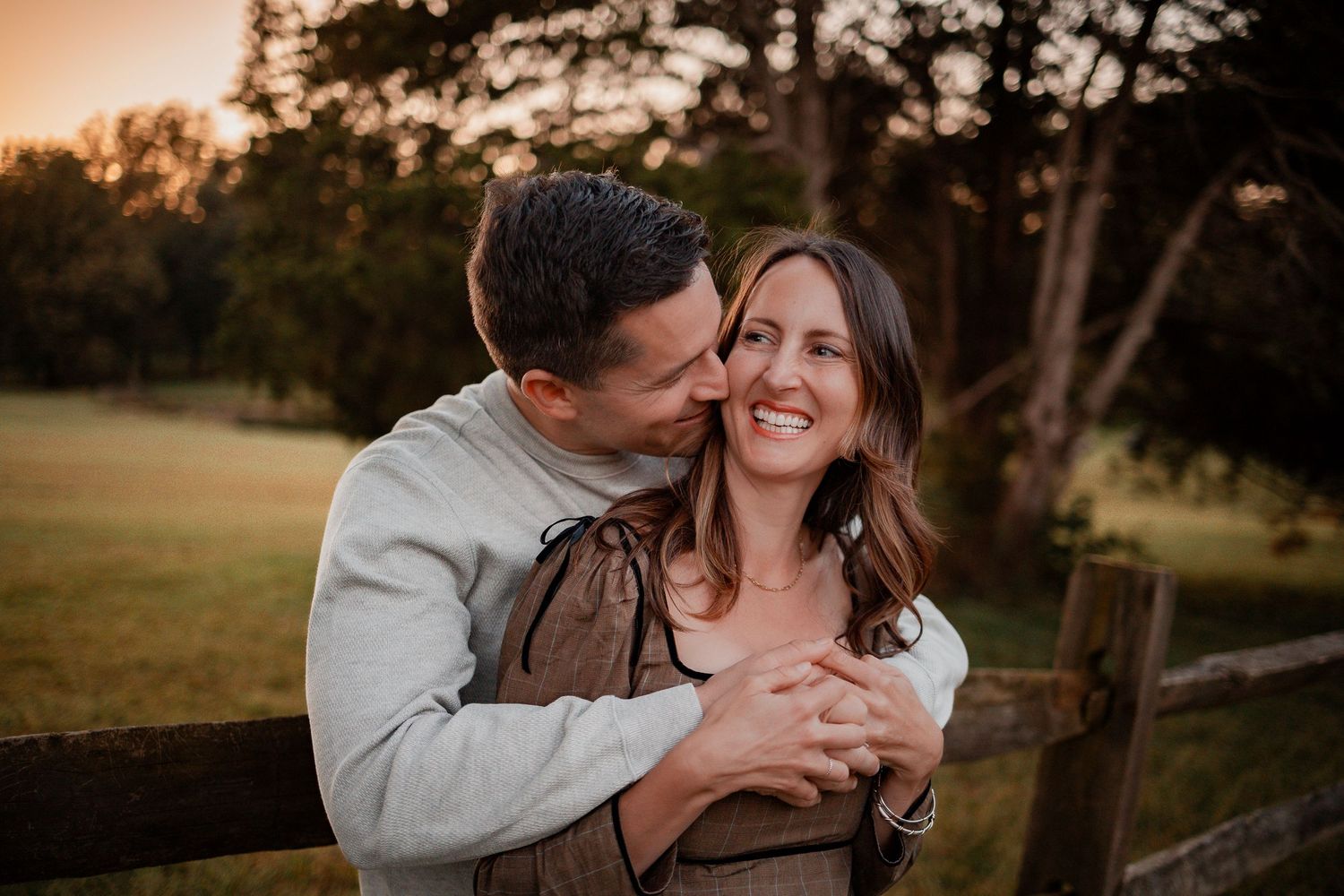 A couple shares an intimate embrace while leaning against a wooden fence at golden hour.