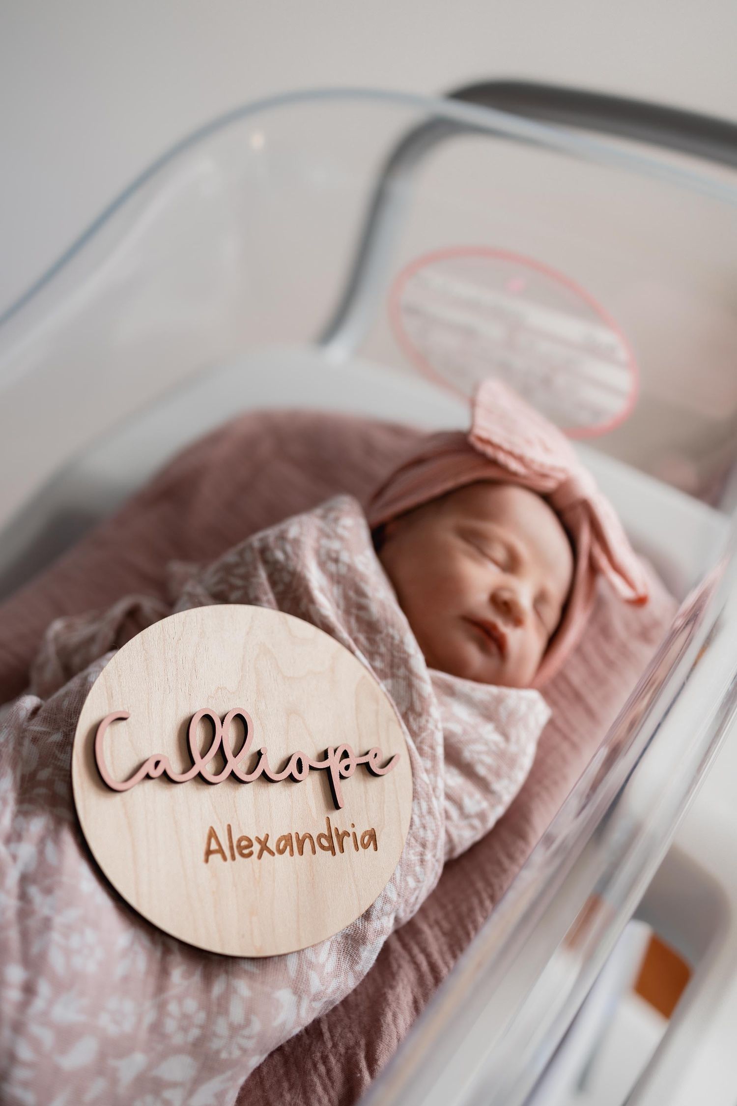 Newborn baby girl named Calliope Alexandria sleeping in hospital bassinet with pink bow headband and floral swaddle.