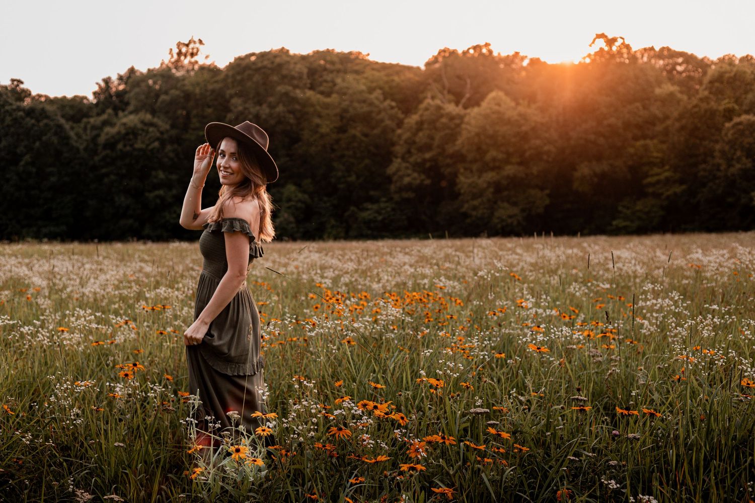 A couple embraces in a wildflower meadow at sunset with golden light filtering through distant trees.