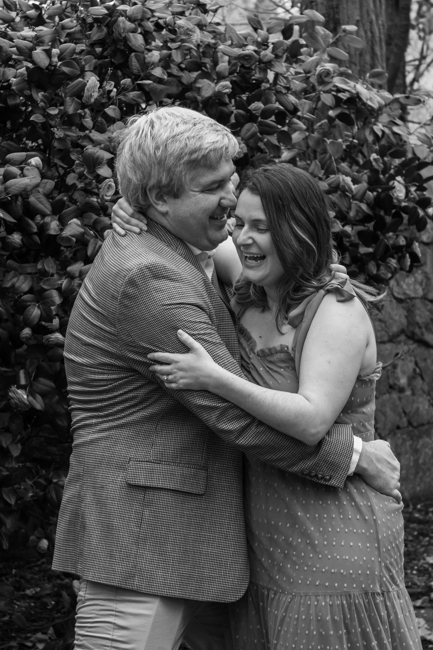 A joyful black and white photo of a couple embracing and laughing together against a leafy background.
