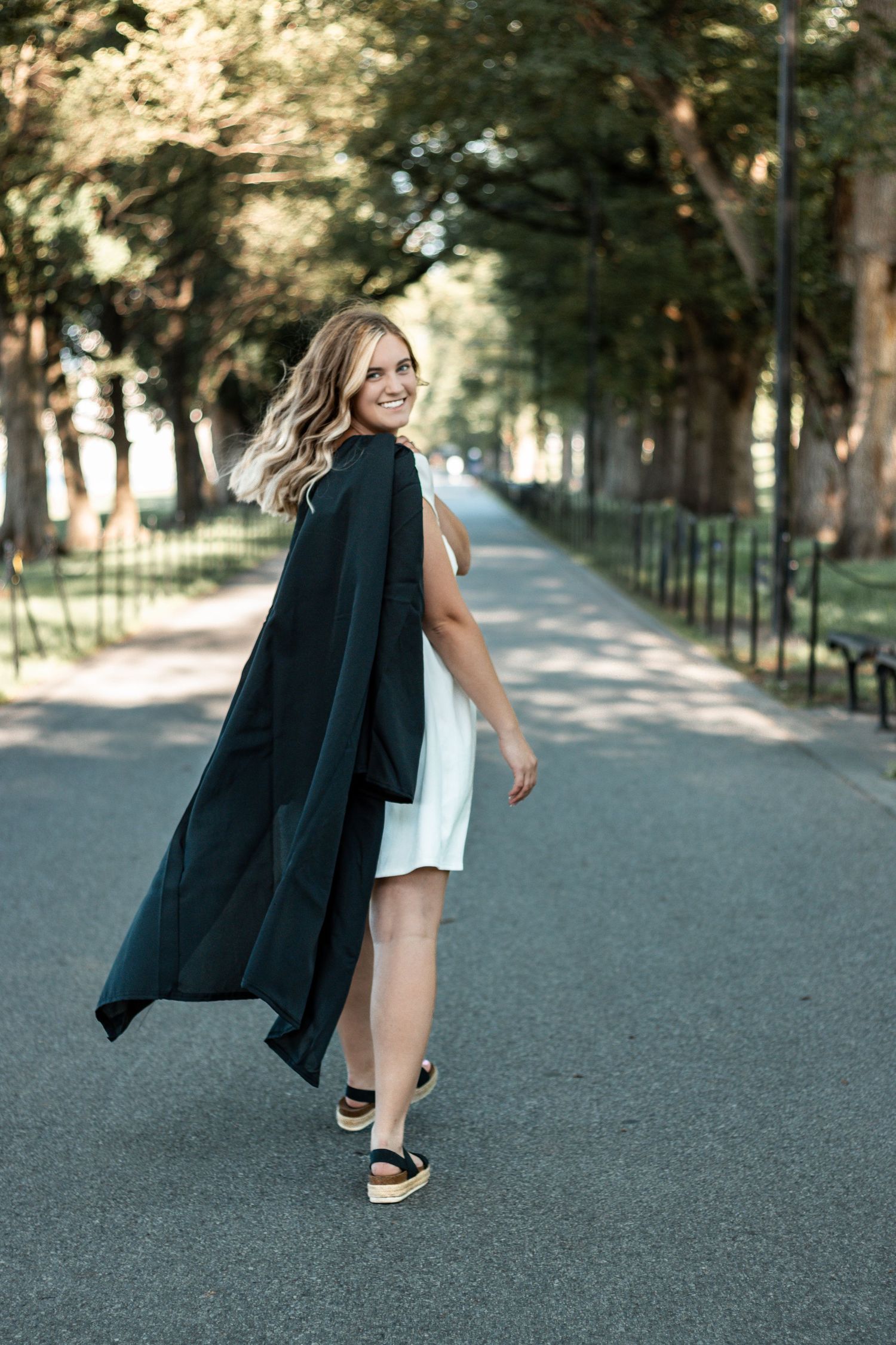 Graduate throws graduation gown while walking along a scenic campus path.