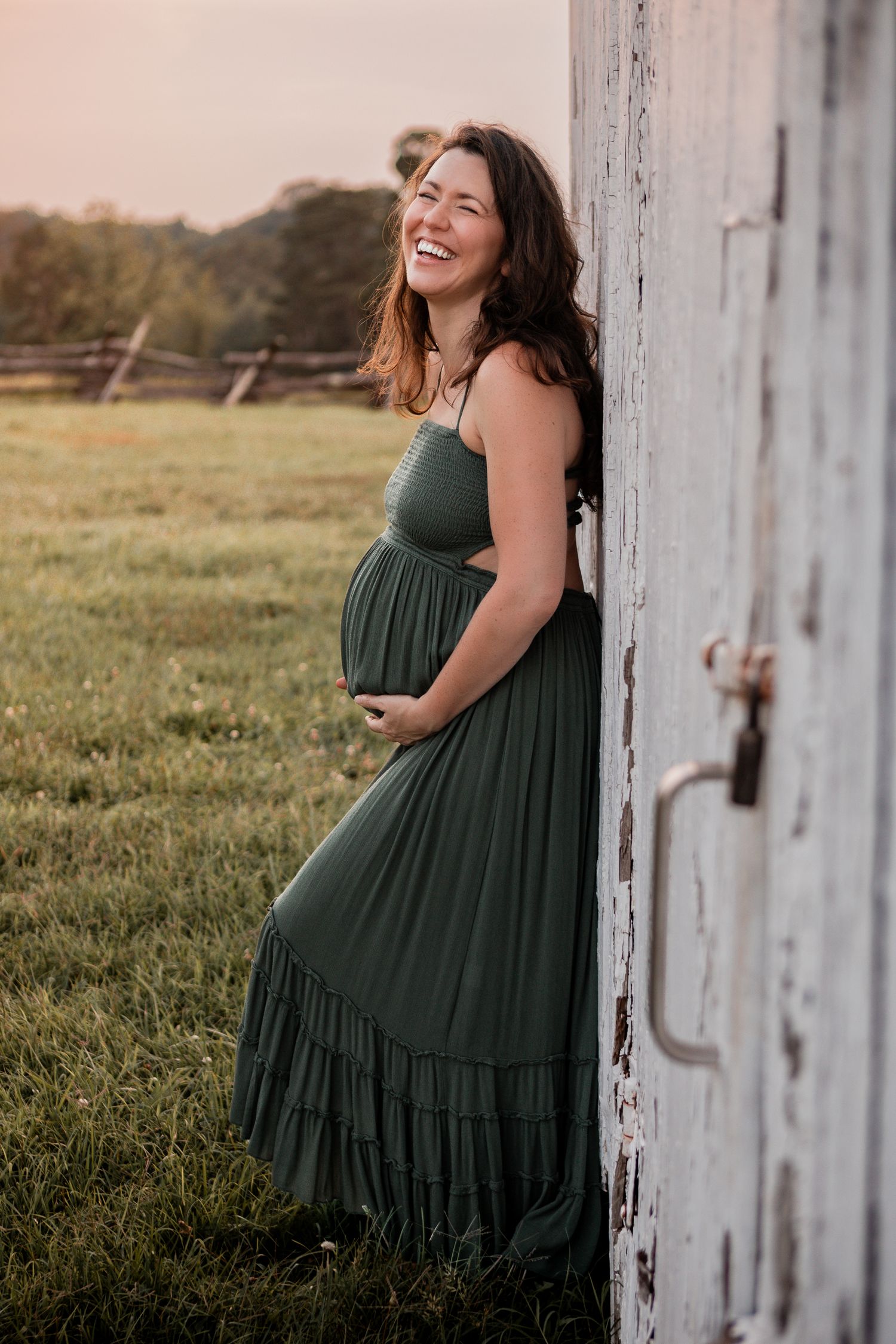 A woman in a dark green maxi dress poses in front of a rustic white barn door during sunset.
