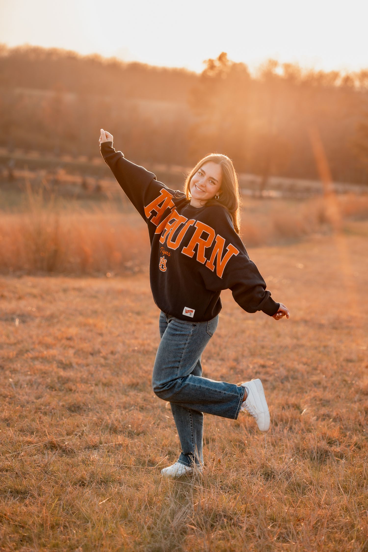 Young woman in Auburn sweatshirt and jeans posing joyfully in golden field at sunset.