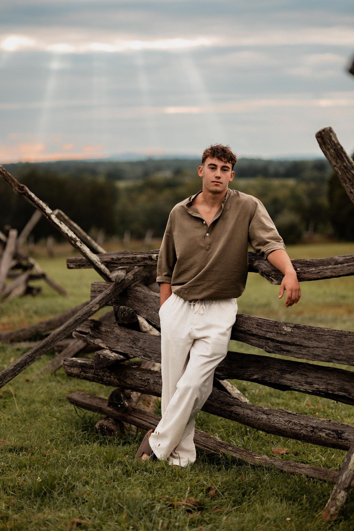 Young man in olive shirt and white pants leaning against rustic wooden fence in green field at sunset.