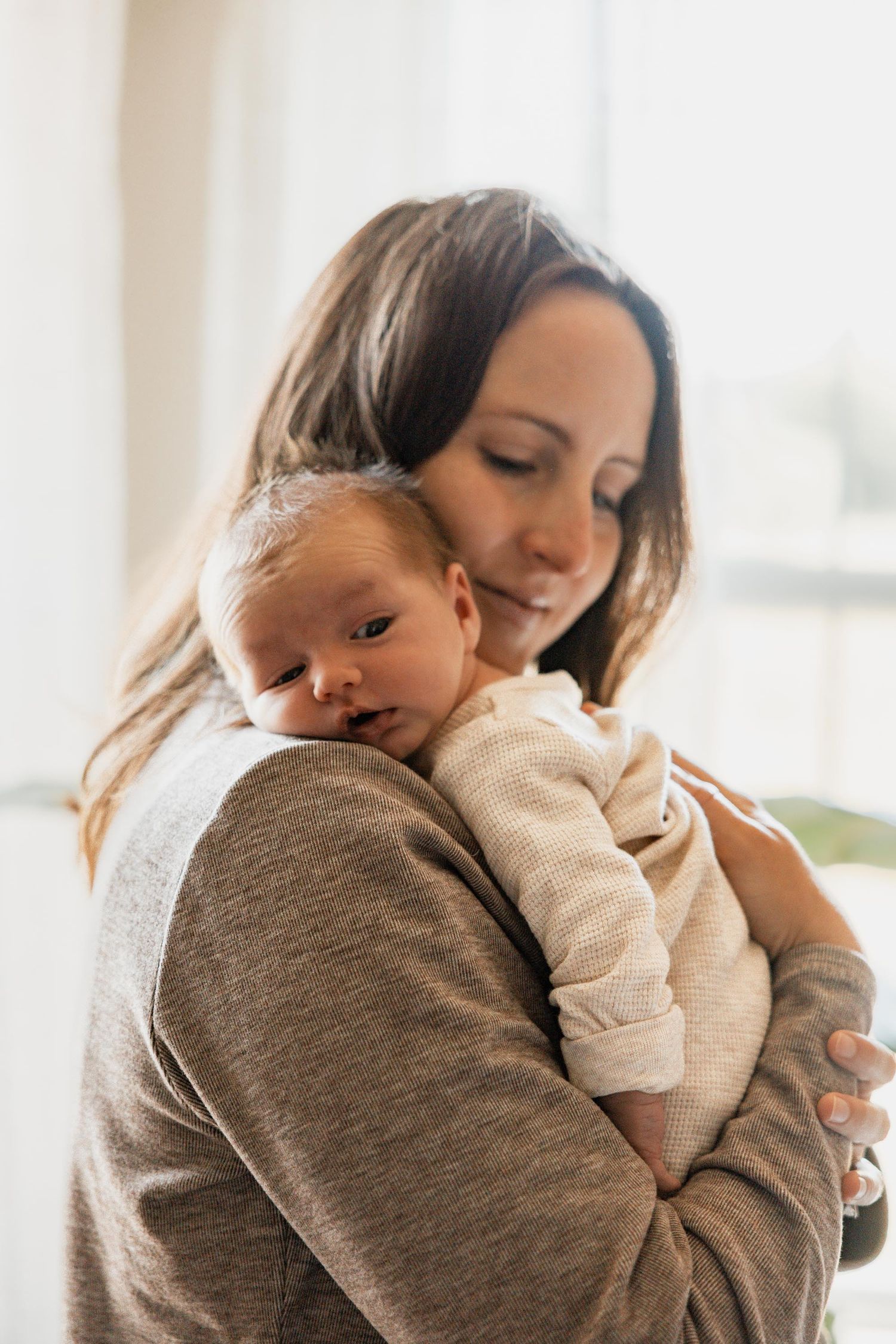 Mother tenderly holding sleeping newborn baby against her chest near a bright window.