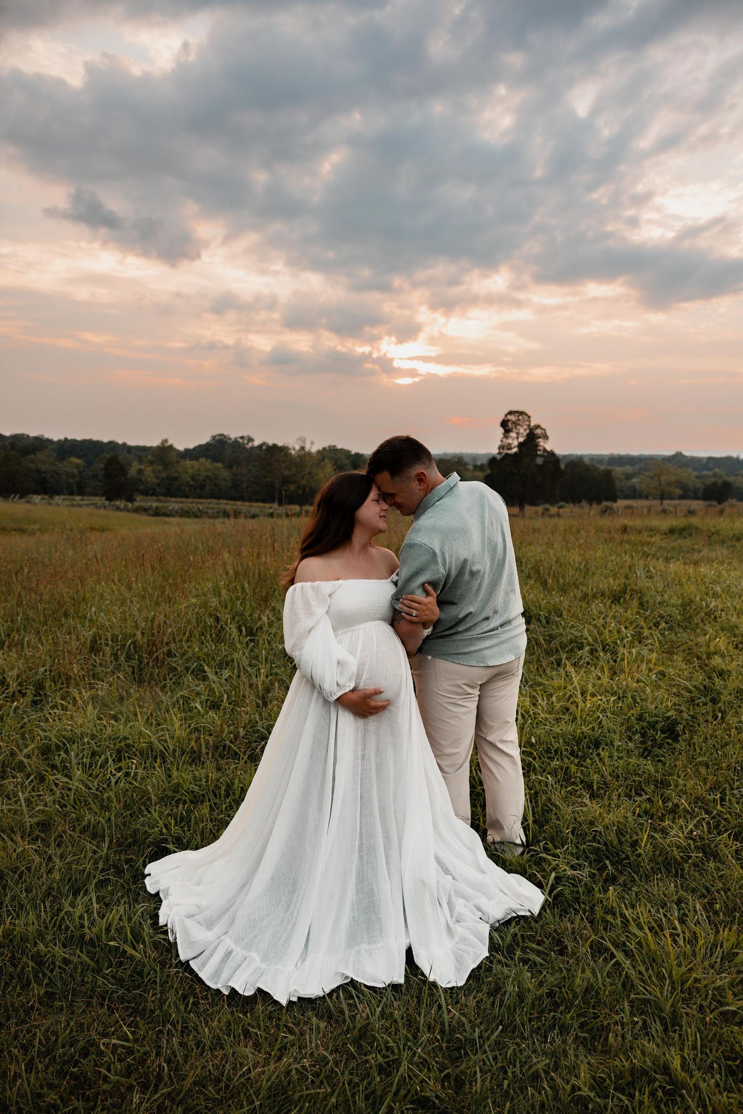 Pregnant couple sharing a tender kiss at sunset in a green field, woman in flowing white off-shoulder gown.
