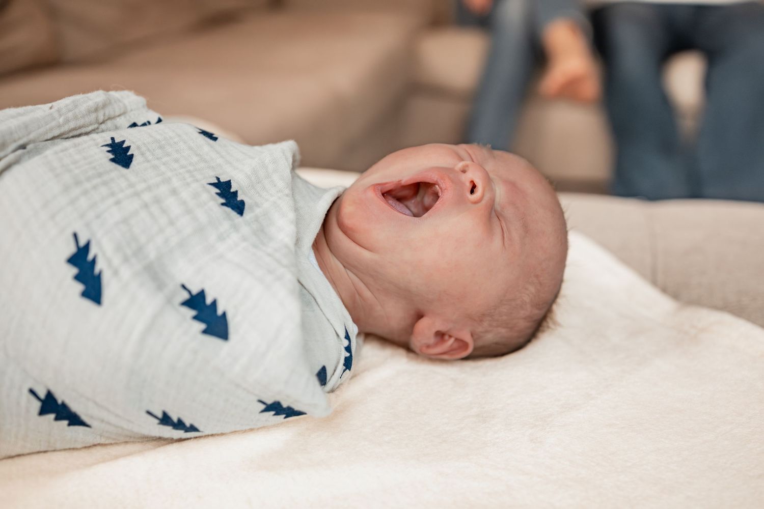 Baby wearing white onesie with blue trees printed on it lies on soft cream colored bedding.