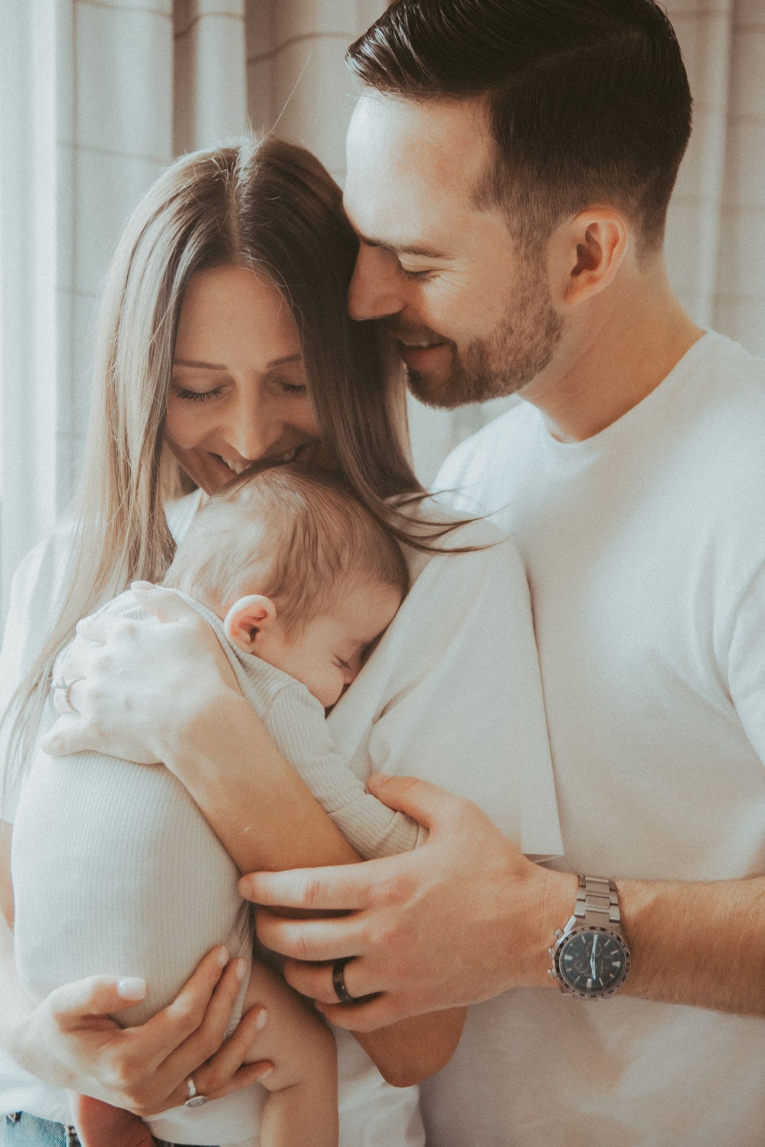 Family portrait series showing parents in white shirts embracing their newborn against a bright window.