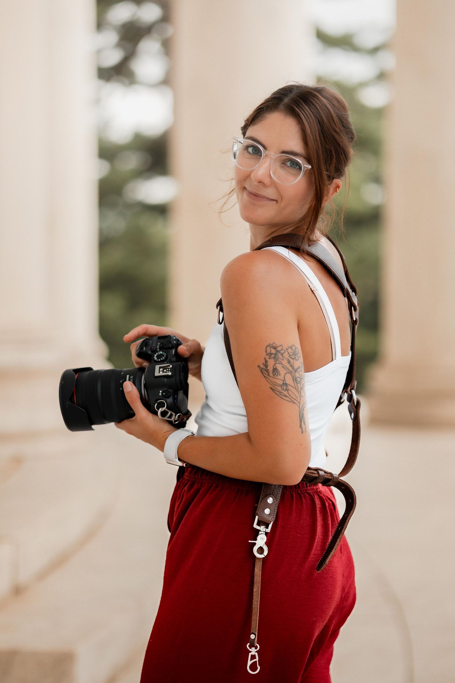 Young woman photographer holding DSLR camera, wearing glasses and red pants, with arm tattoo, smiling outdoors near columns.