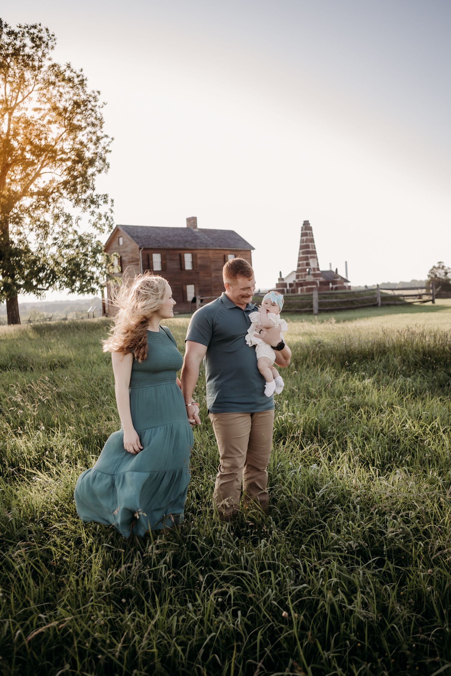 A couple embraces in a grassy field at sunset with a rustic barn in the distant background.
