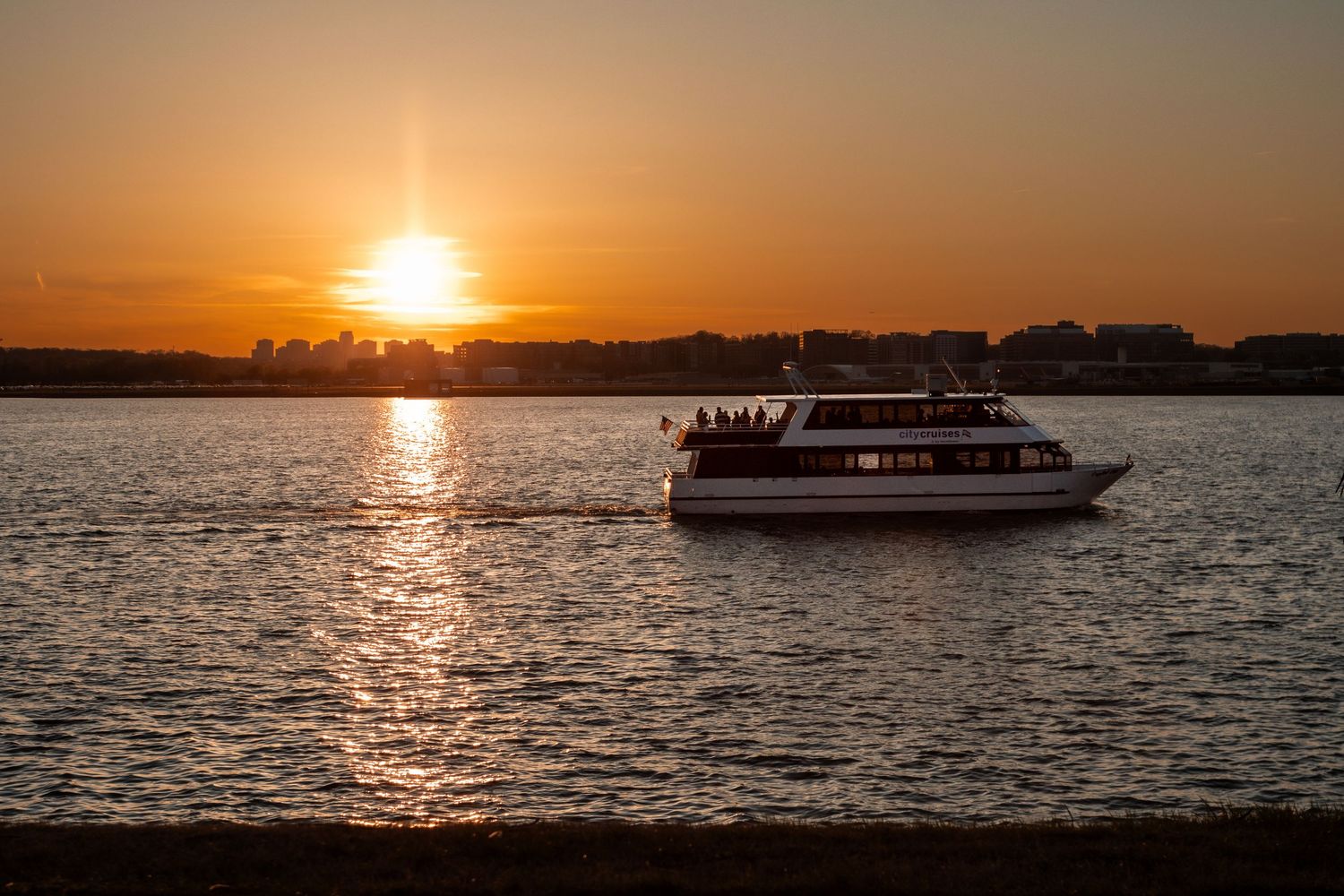 A boat cruises across a lake during a golden sunset with silhouetted trees along the shoreline.