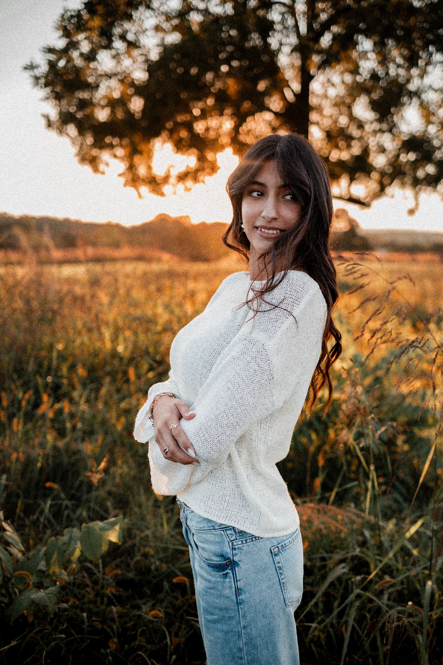 A woman in a white sweater and jeans dances in a golden field at sunset.