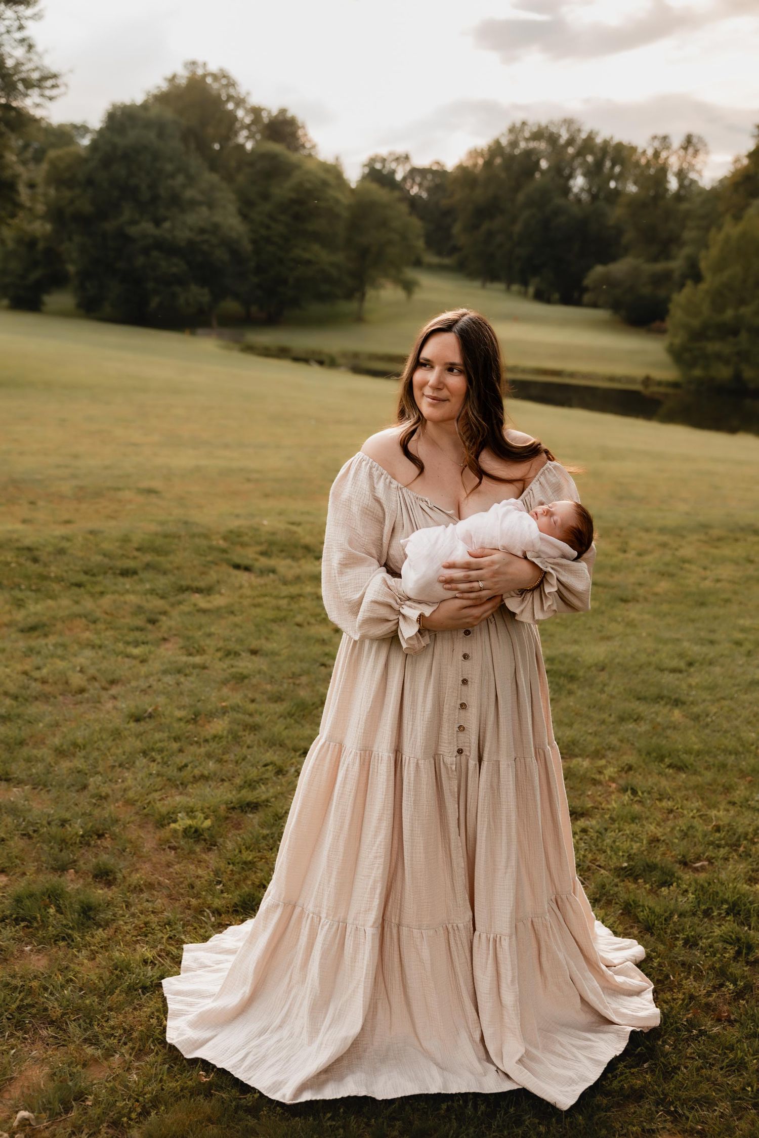A mother in a flowing beige dress holds her newborn baby in a lush green field at golden hour.