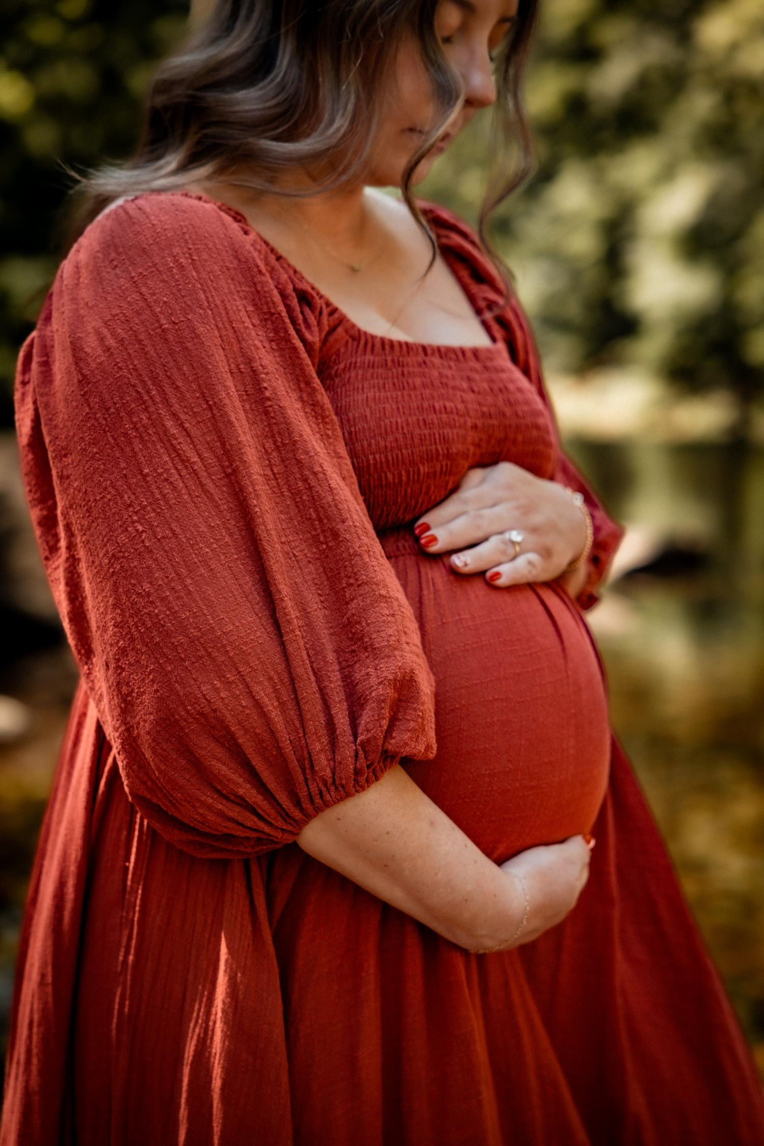 Pregnant woman in rust red smocked dress cradling baby bump outdoors in natural setting.