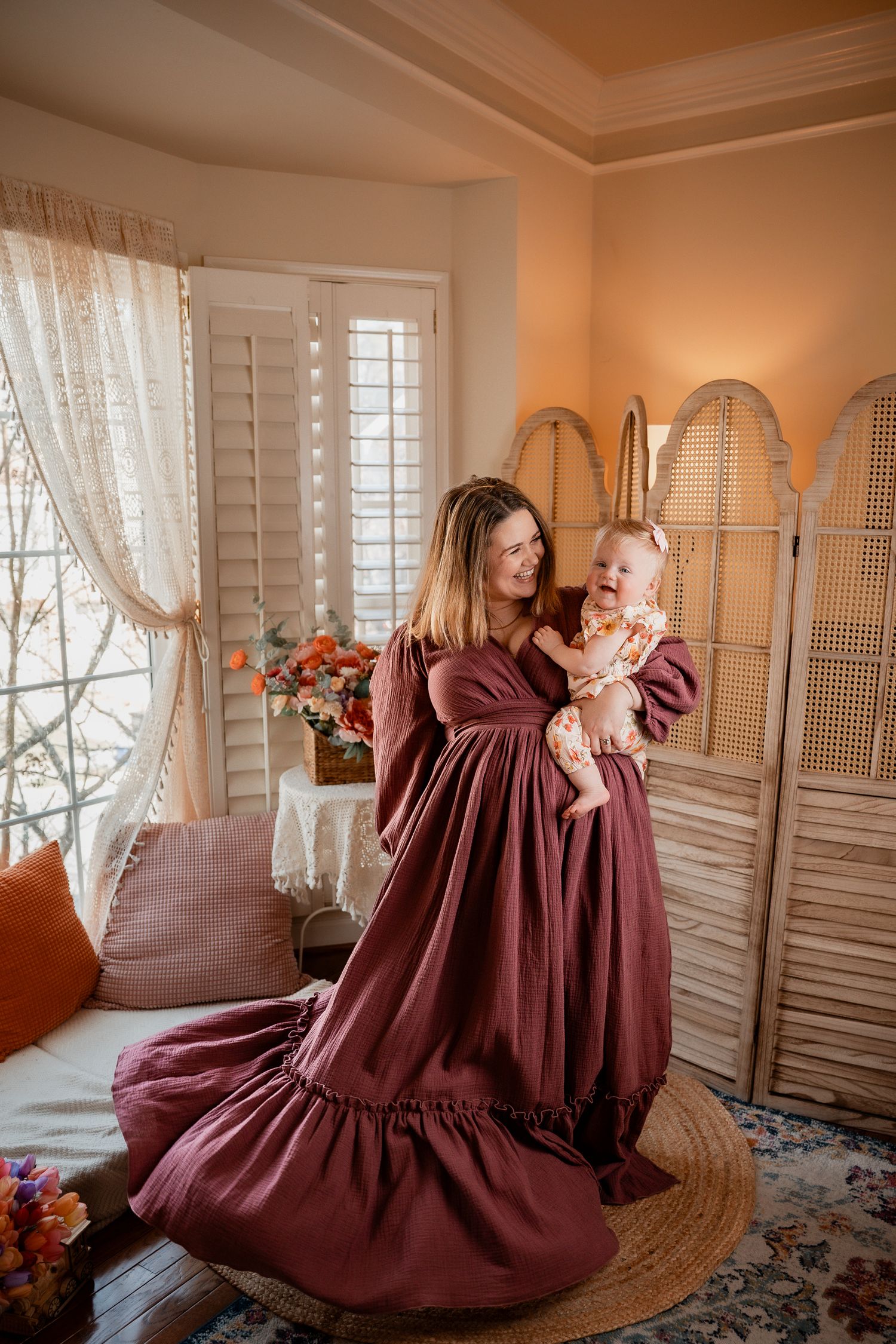 A mother in a mauve dress holds her baby in a warmly lit room with shuttered windows and a rattan screen.
