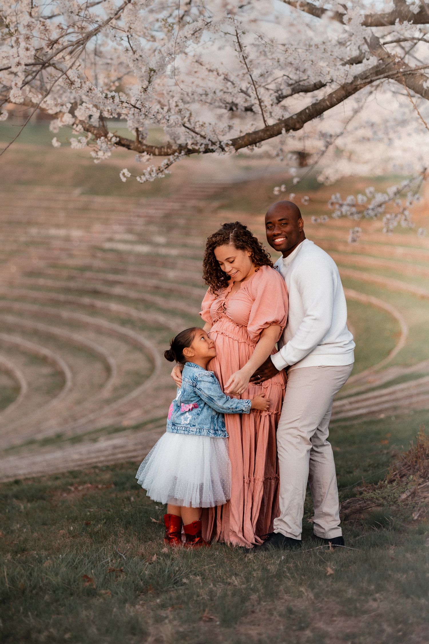A family maternity photo under cherry blossoms, with a pregnant woman in pink, her partner, and young daughter in a tutu.