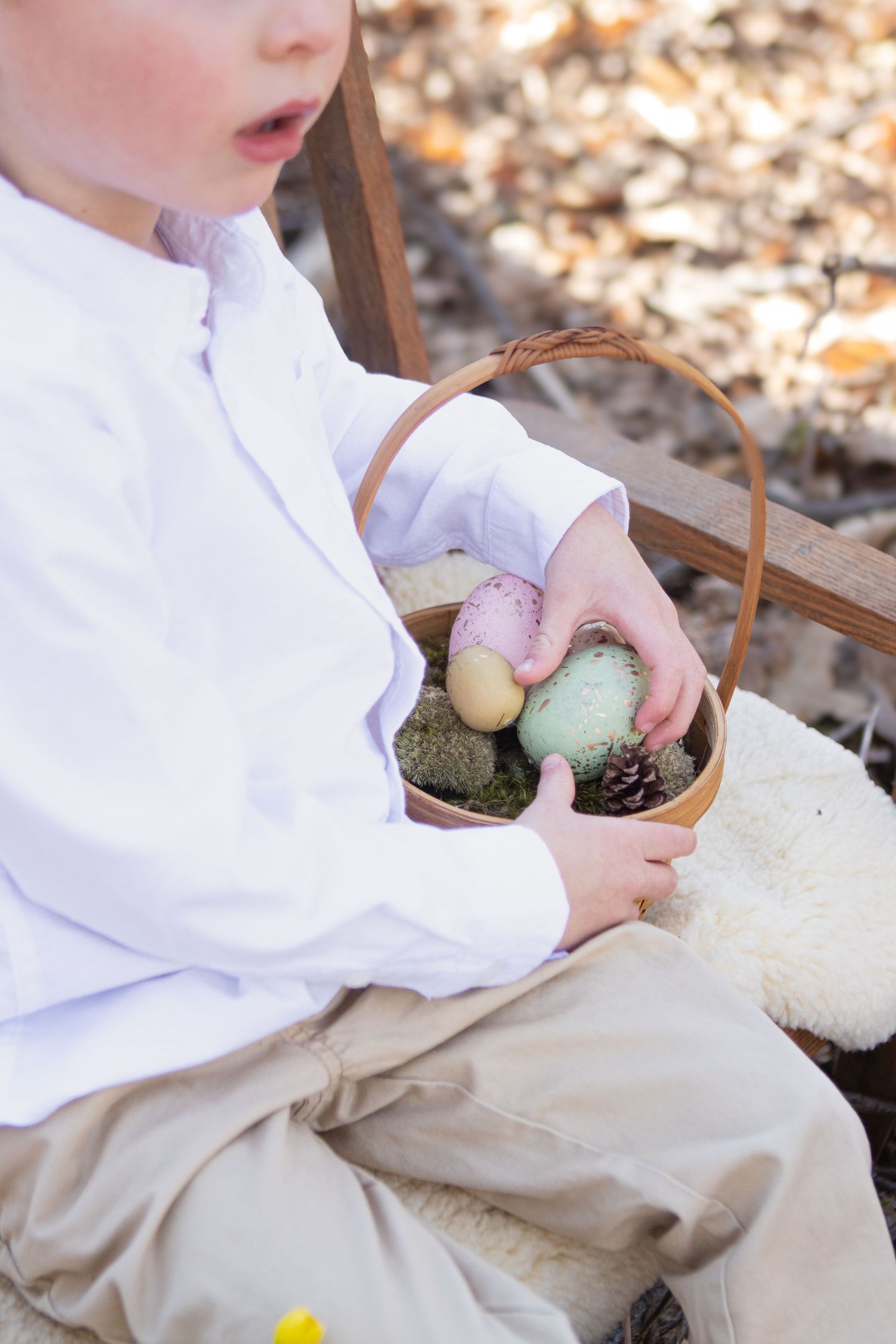 Child in white shirt holds wicker basket with colorful Easter eggs and moss.