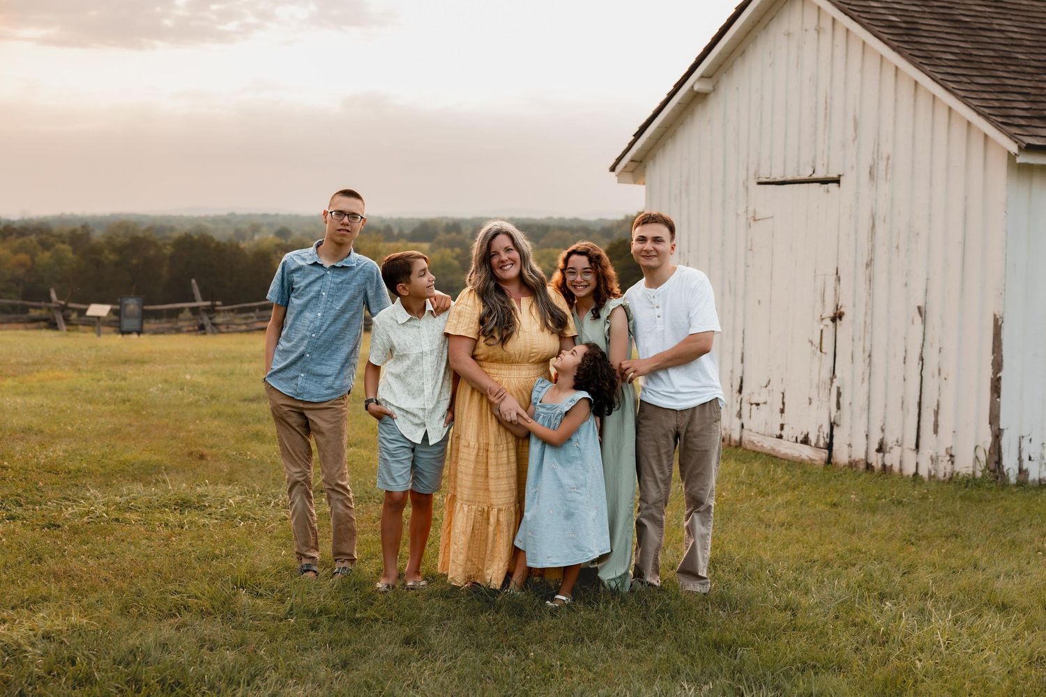 Happy family of five poses together smiling near a white barn at golden hour.