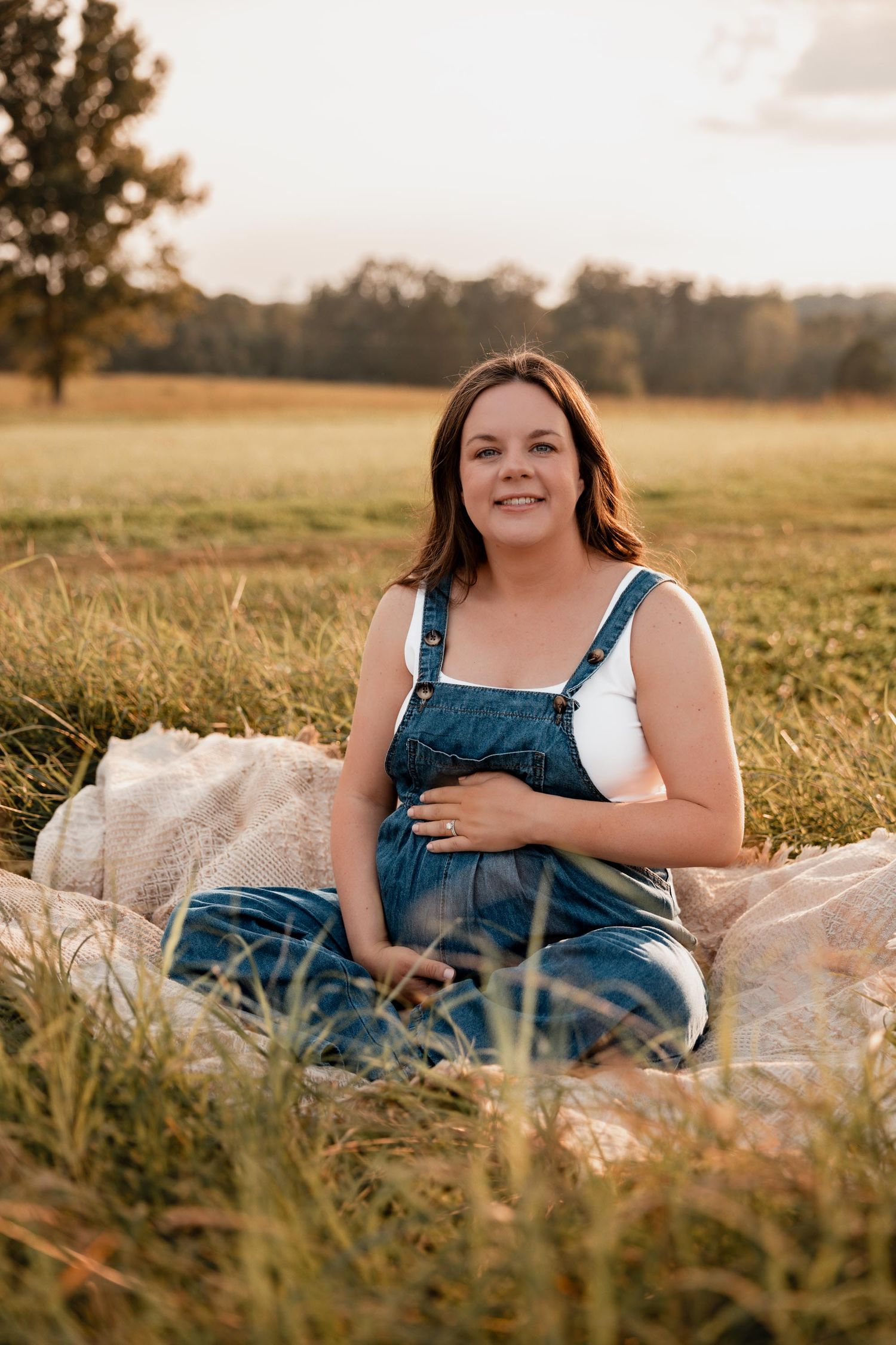 Smiling pregnant woman in denim overalls sitting on blanket in golden hour field, gently holding her baby bump.