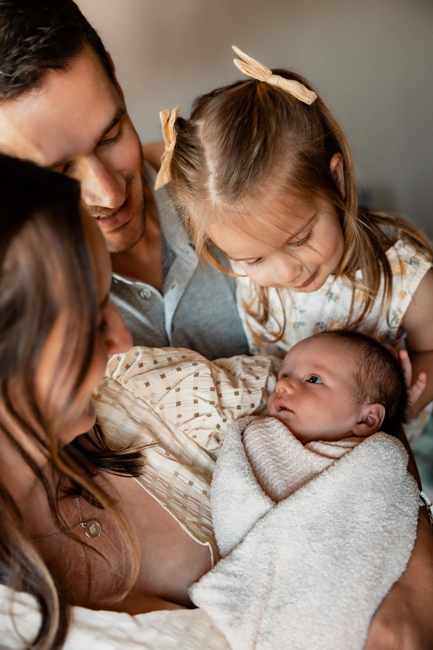 Family of four admiring their newborn baby, with parents and toddler girl gathering closely together in a warm, tender moment.