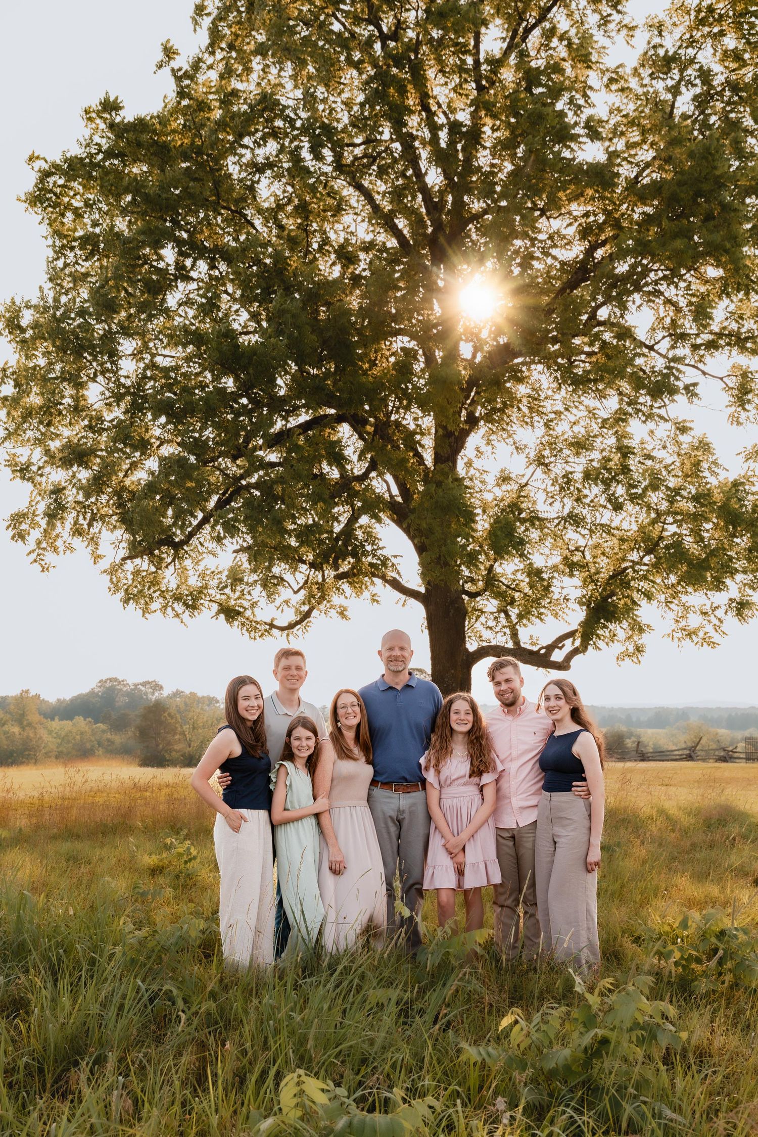 A large family of eight poses together in a sunlit field beneath a sprawling oak tree during golden hour.