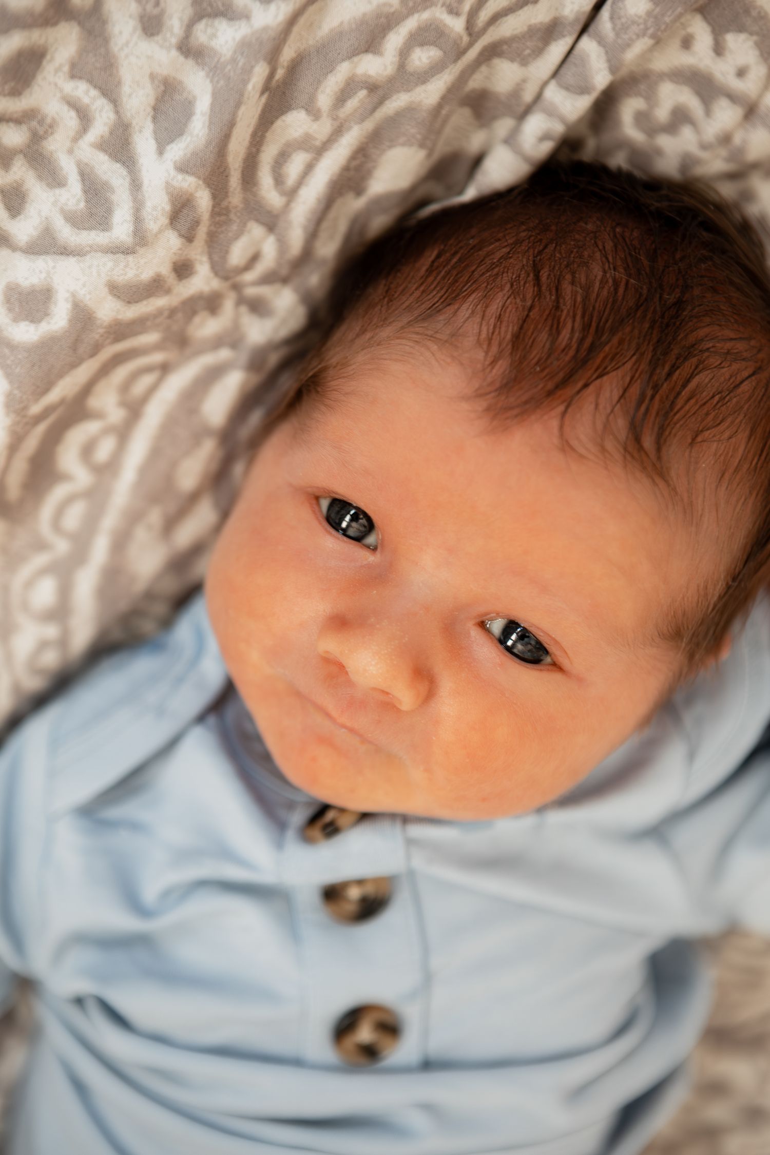 A close-up portrait of a newborn baby wearing a light blue outfit with buttons.