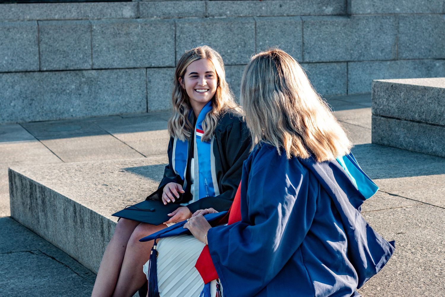 A graduate takes joyful photos in a navy blue cap and gown with red stole outside a historic building.