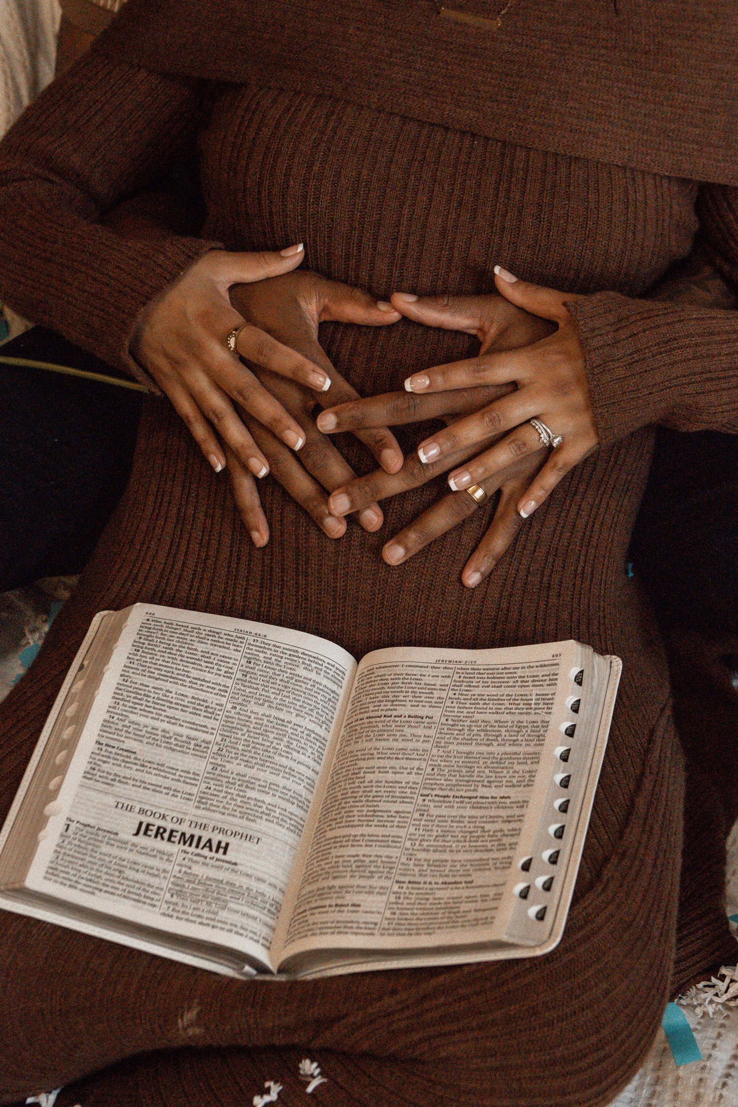 Hands rest on a brown sweater above an open Bible displaying the book of Jeremiah.
