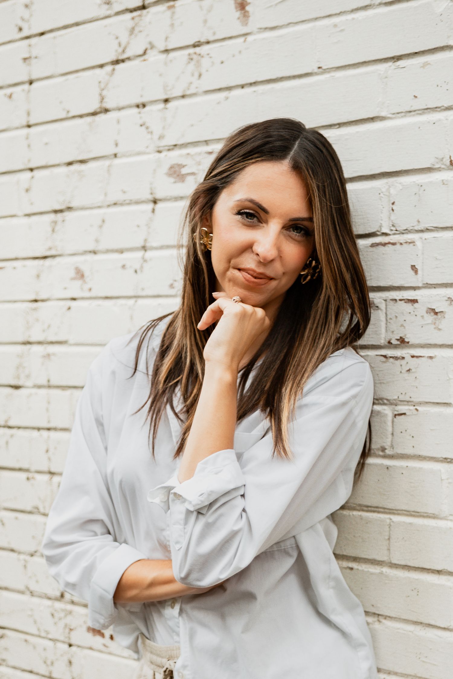 Casual portrait of someone with long brown hair wearing a white button-down shirt leaning against a white painted wall.