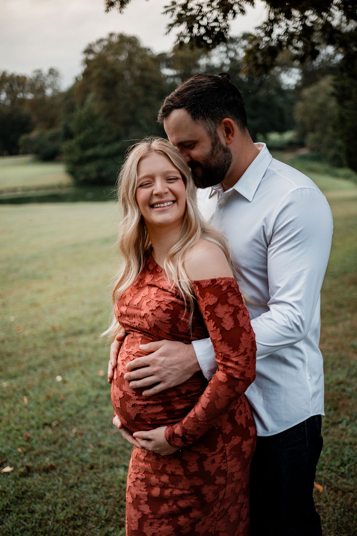 Couple embraces in a grassy field during maternity photoshoot wearing rust dress and white shirt.