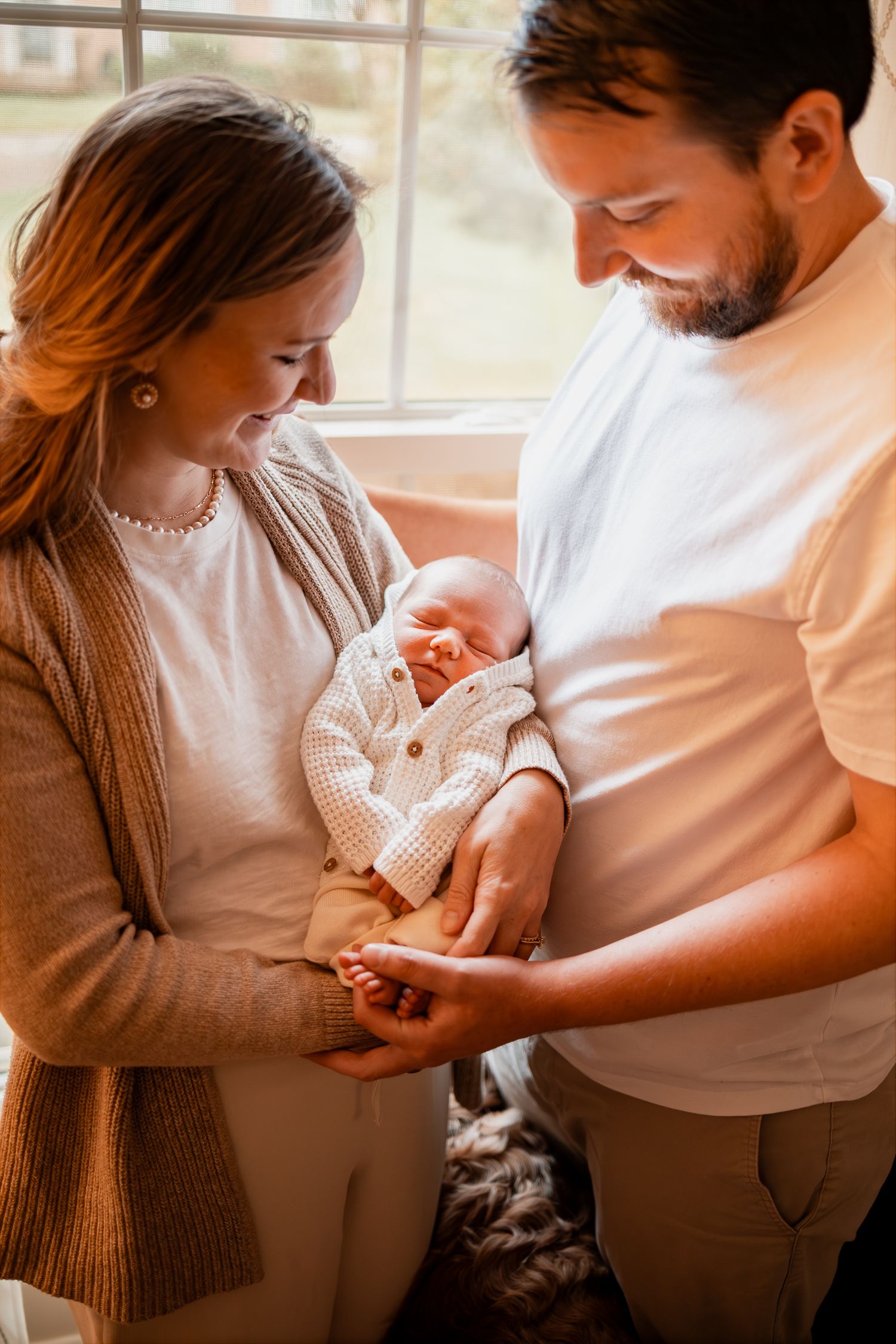 Parents holding their newborn baby near a sunlit window in warm, intimate moment.