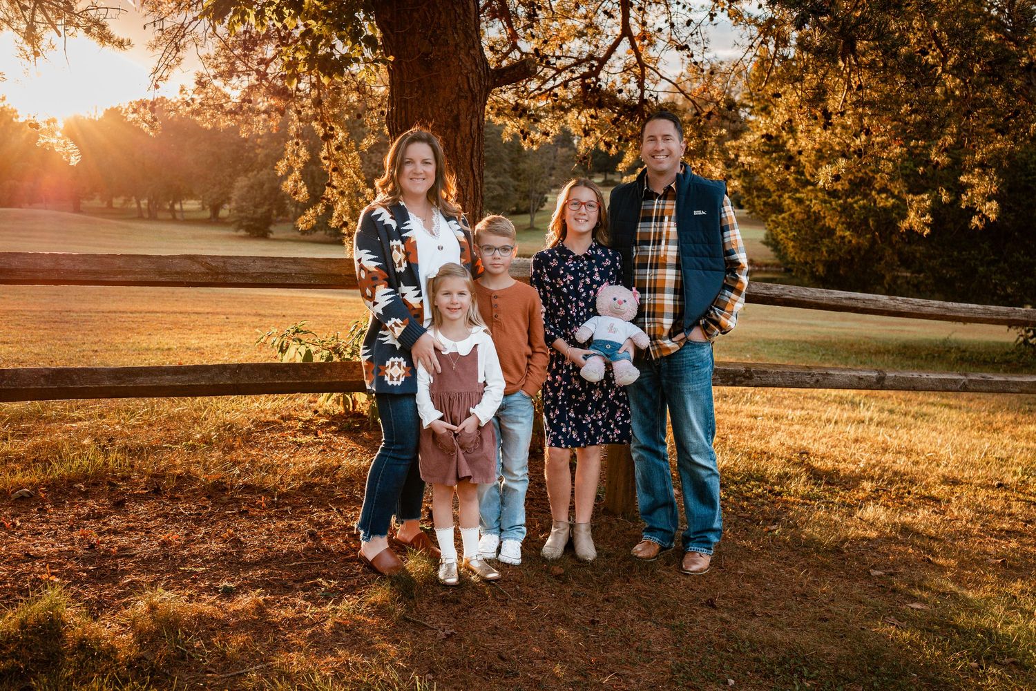 A family poses together under a large tree during golden hour sunset in a grassy park setting.