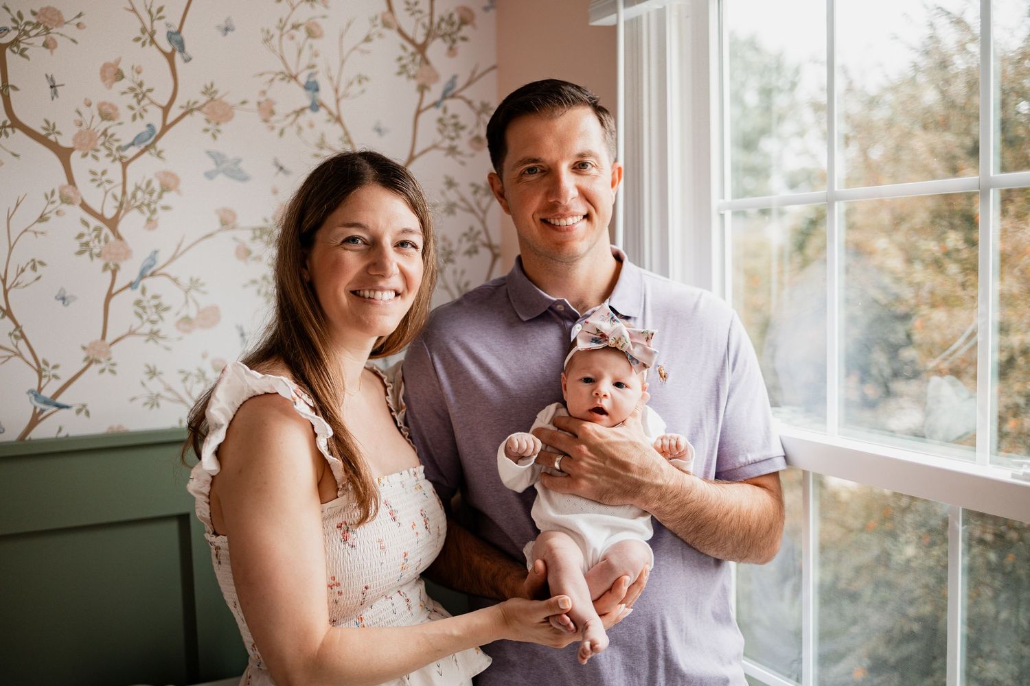 A couple sits together in a bright room with floral wallpaper while holding their newborn baby.