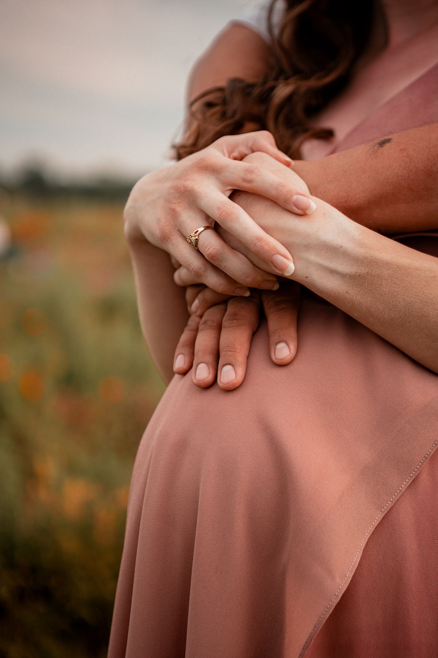 Close-up of hands resting gently on rose-colored fabric with a meadow in soft focus background.