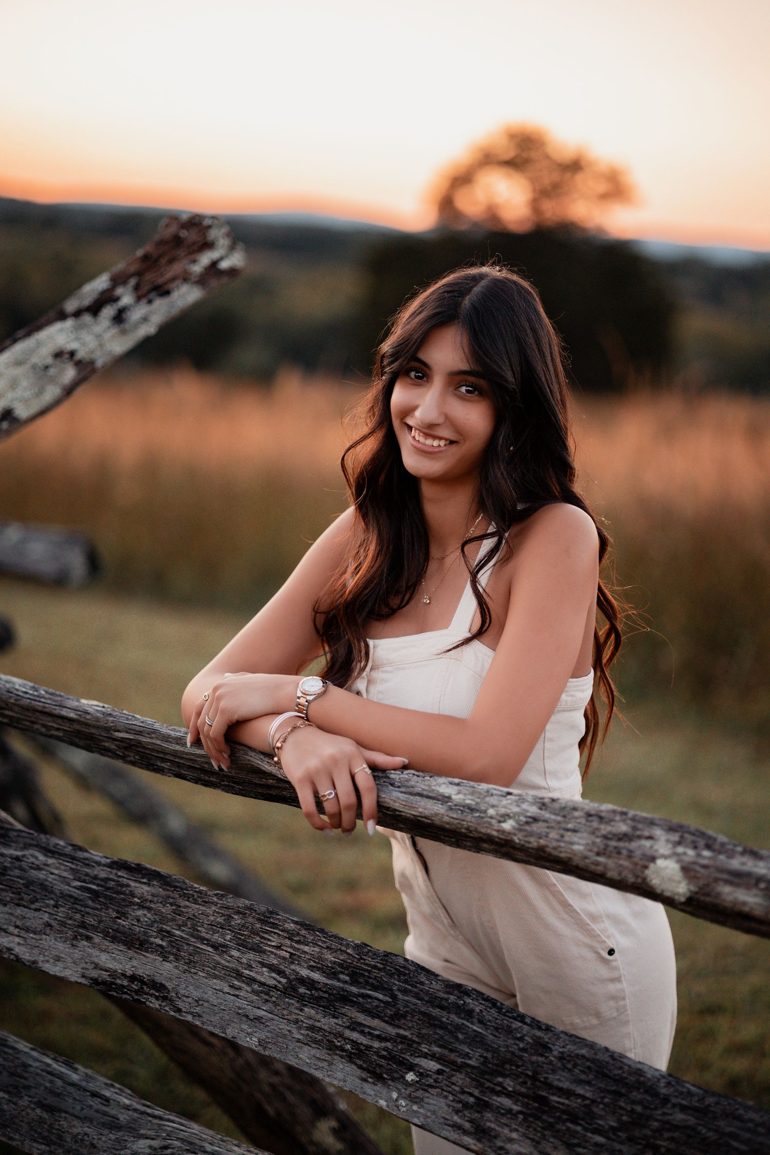 Portrait of a dark-haired woman in white dress leaning on rustic wooden fence at sunset.