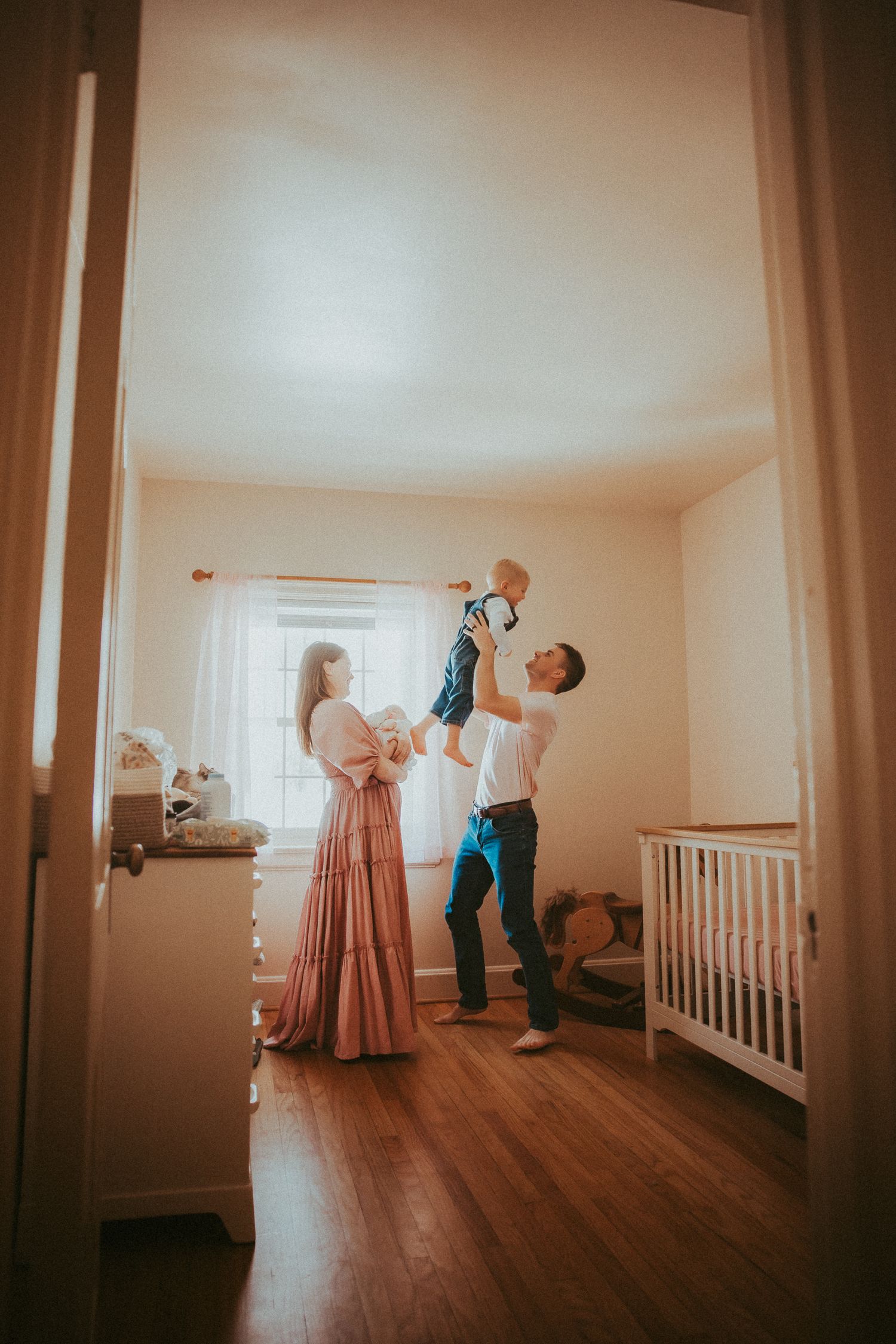 Parents joyfully lifting baby in a cozy nursery with white crib, hardwood floors, and soft natural light.