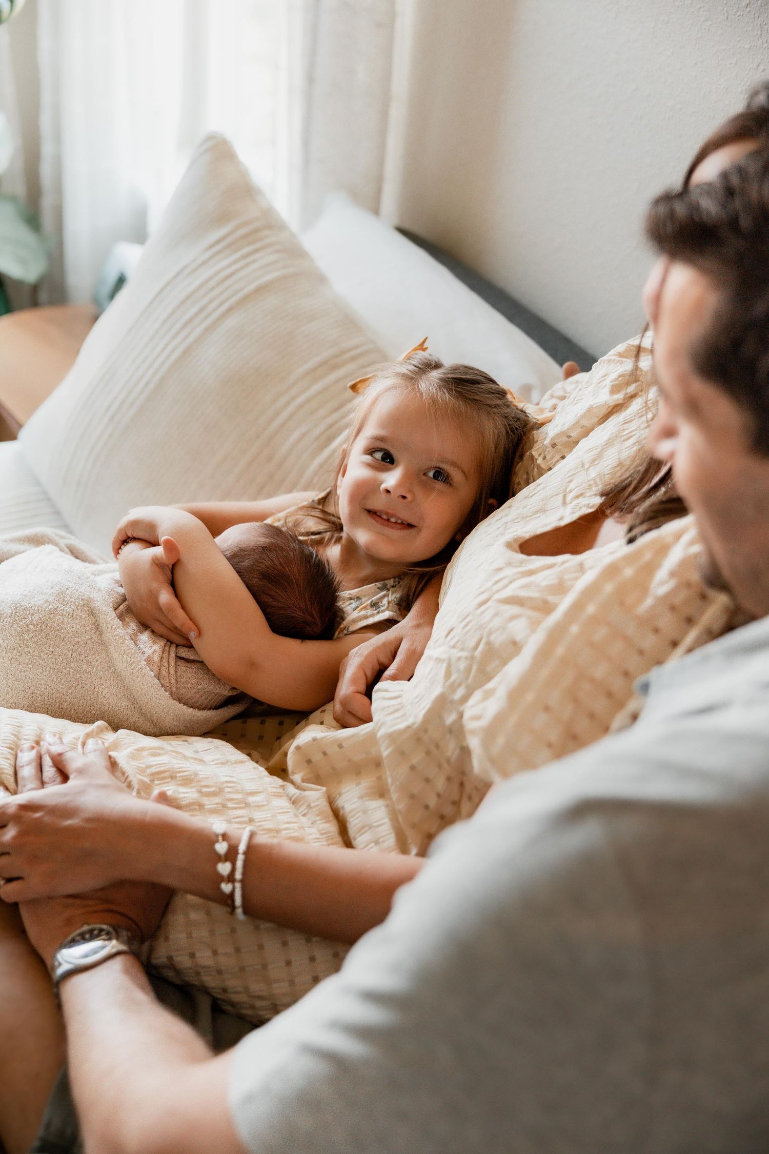 A smiling toddler girl holds a newborn baby on a cozy sofa while a parent watches nearby.