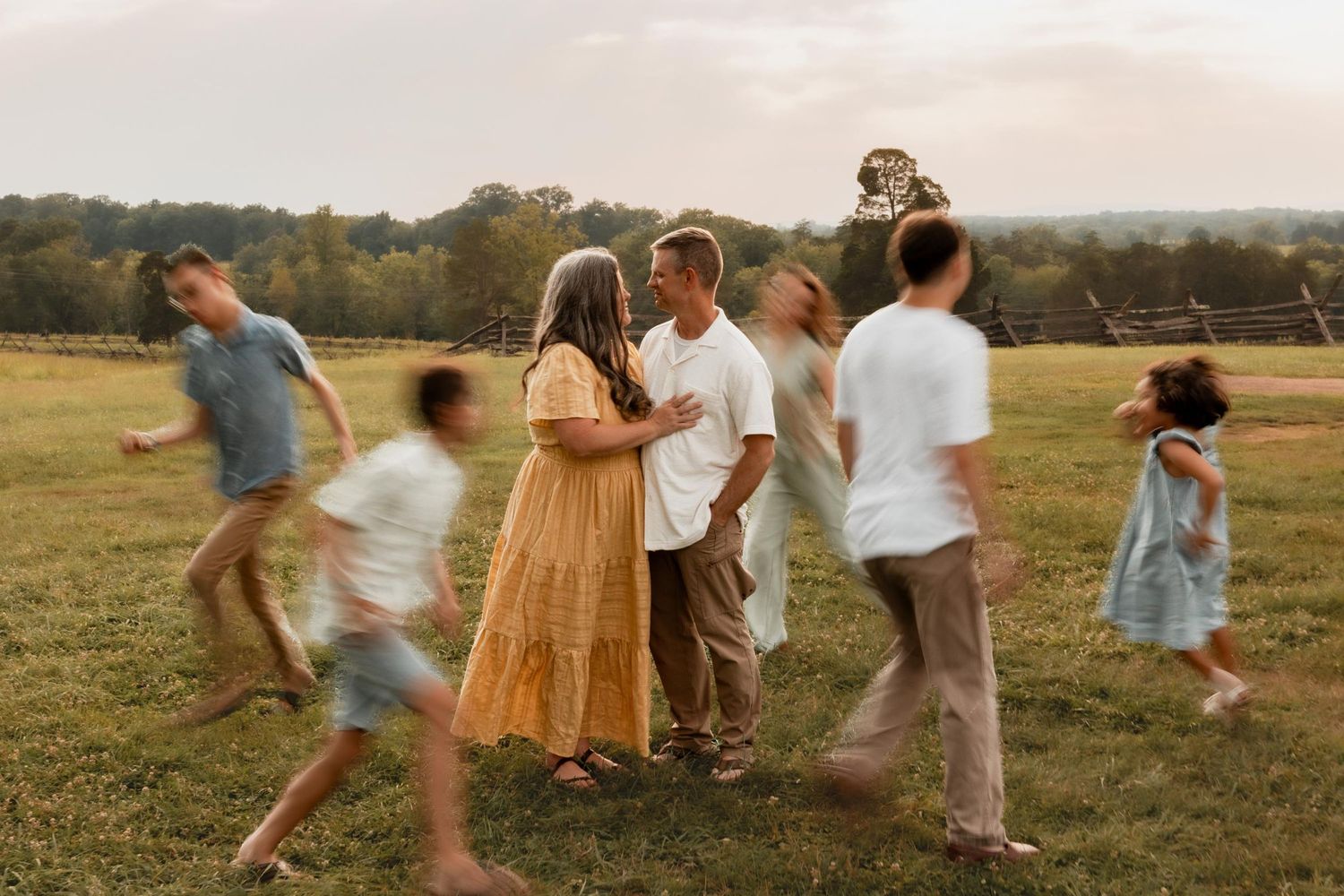 Blurred family members move around a couple embracing in a golden field at sunset.